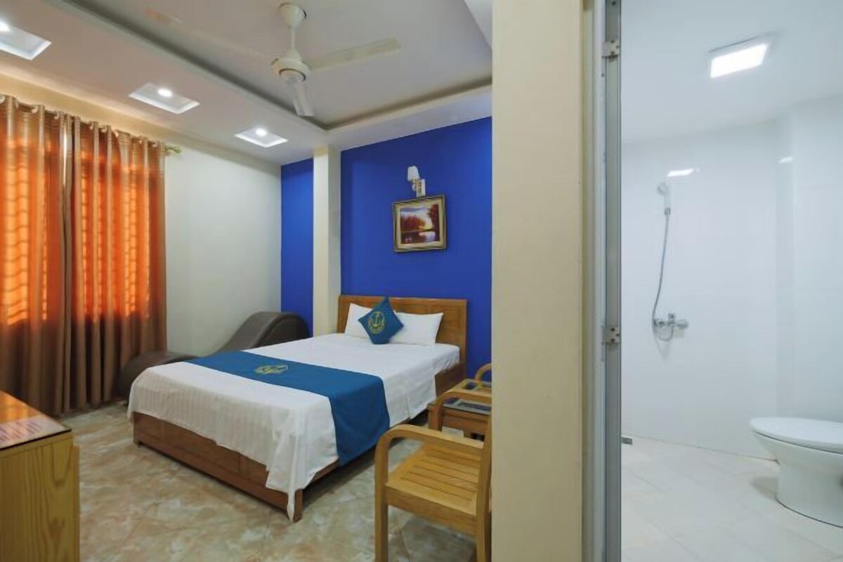 Quarto de hotel com cama, escrivaninha, cadeira, chuveiro e vaso sanitário.