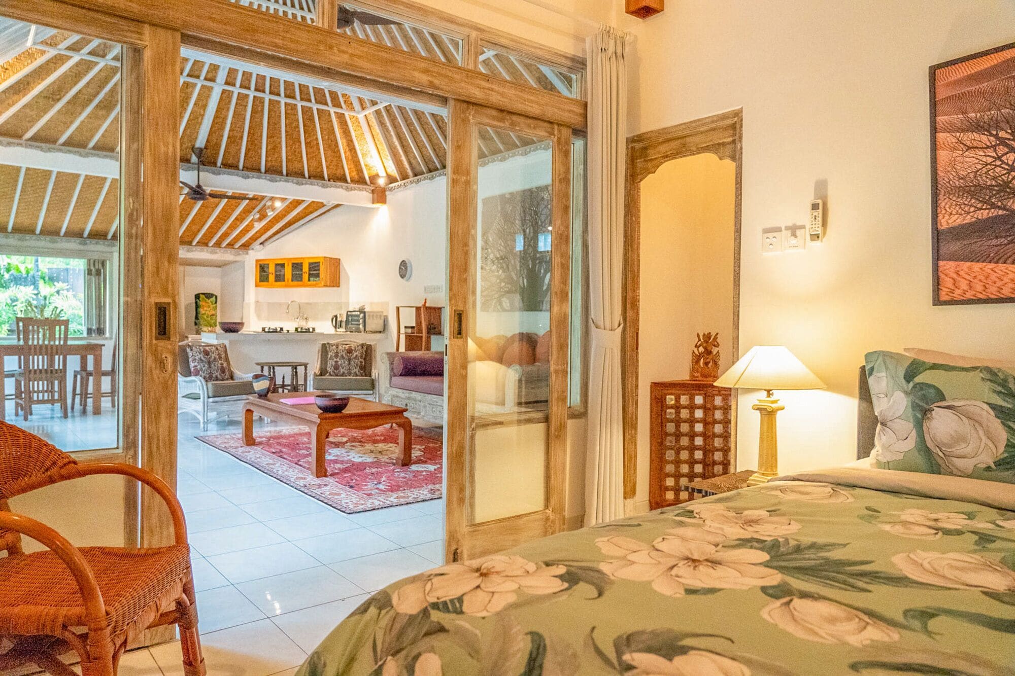 A room with a bed, a chair, a table, and a view of a kitchen and dining area through glass doors.