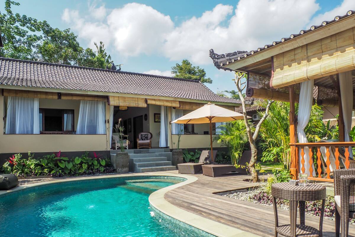 Uma piscina com deck de madeira, espreguiçadeiras e uma área coberta com mesa e guarda-sol.