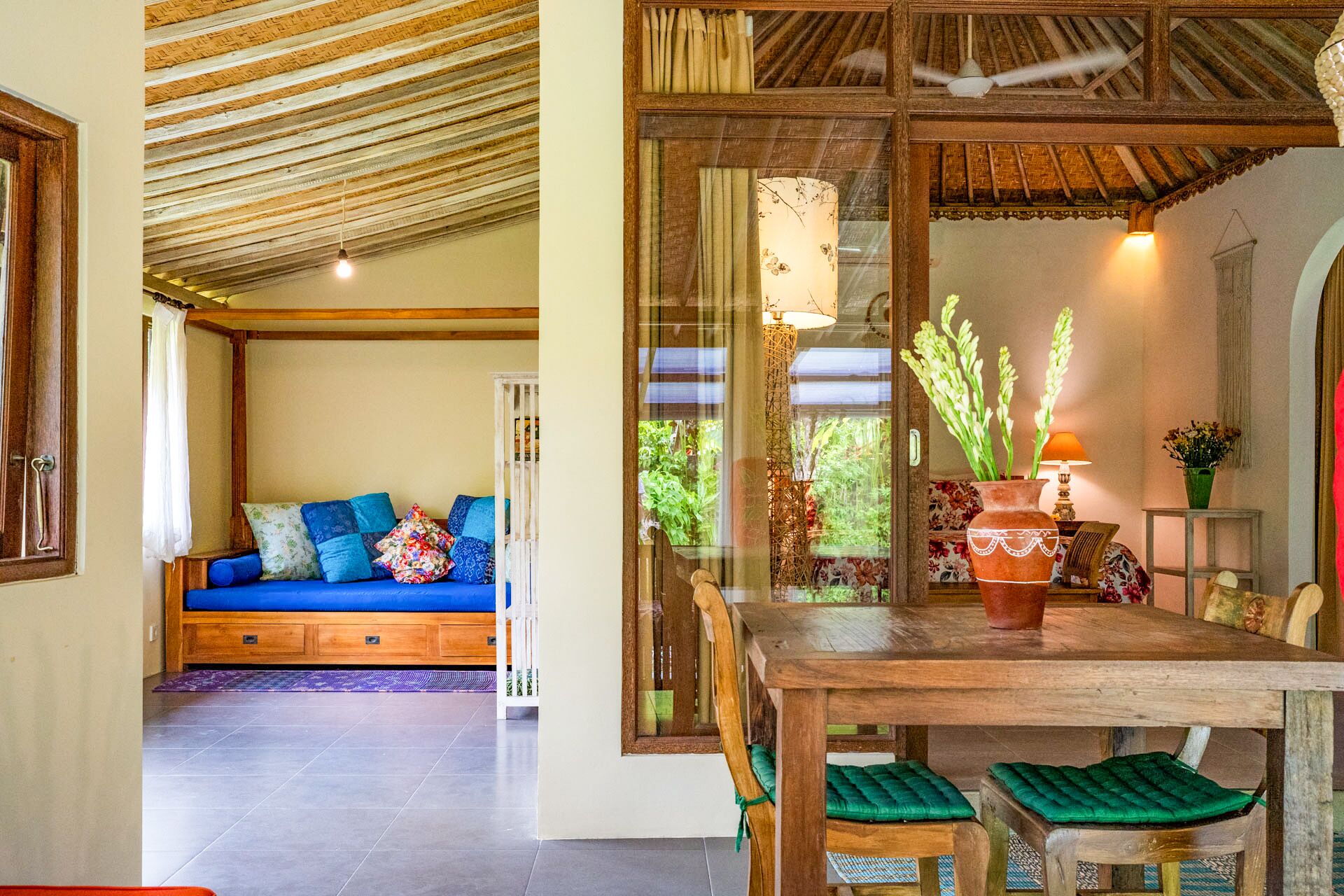 A room with a wooden ceiling, a blue sofa with patterned cushions, a dining table with chairs, and a view of greenery outside.