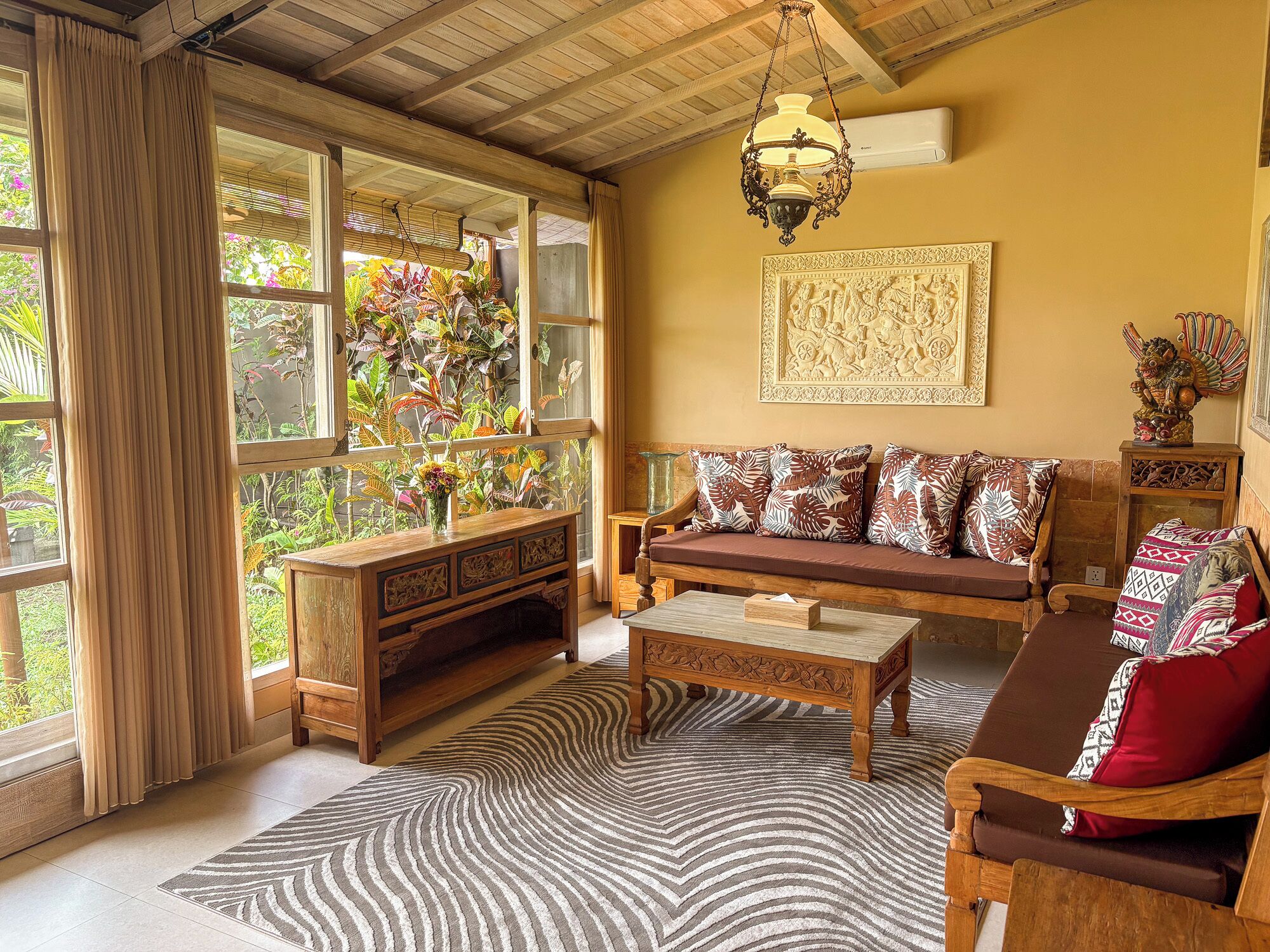 A cozy living room with a wooden sofa, a coffee table, and a large window with a view of greenery.