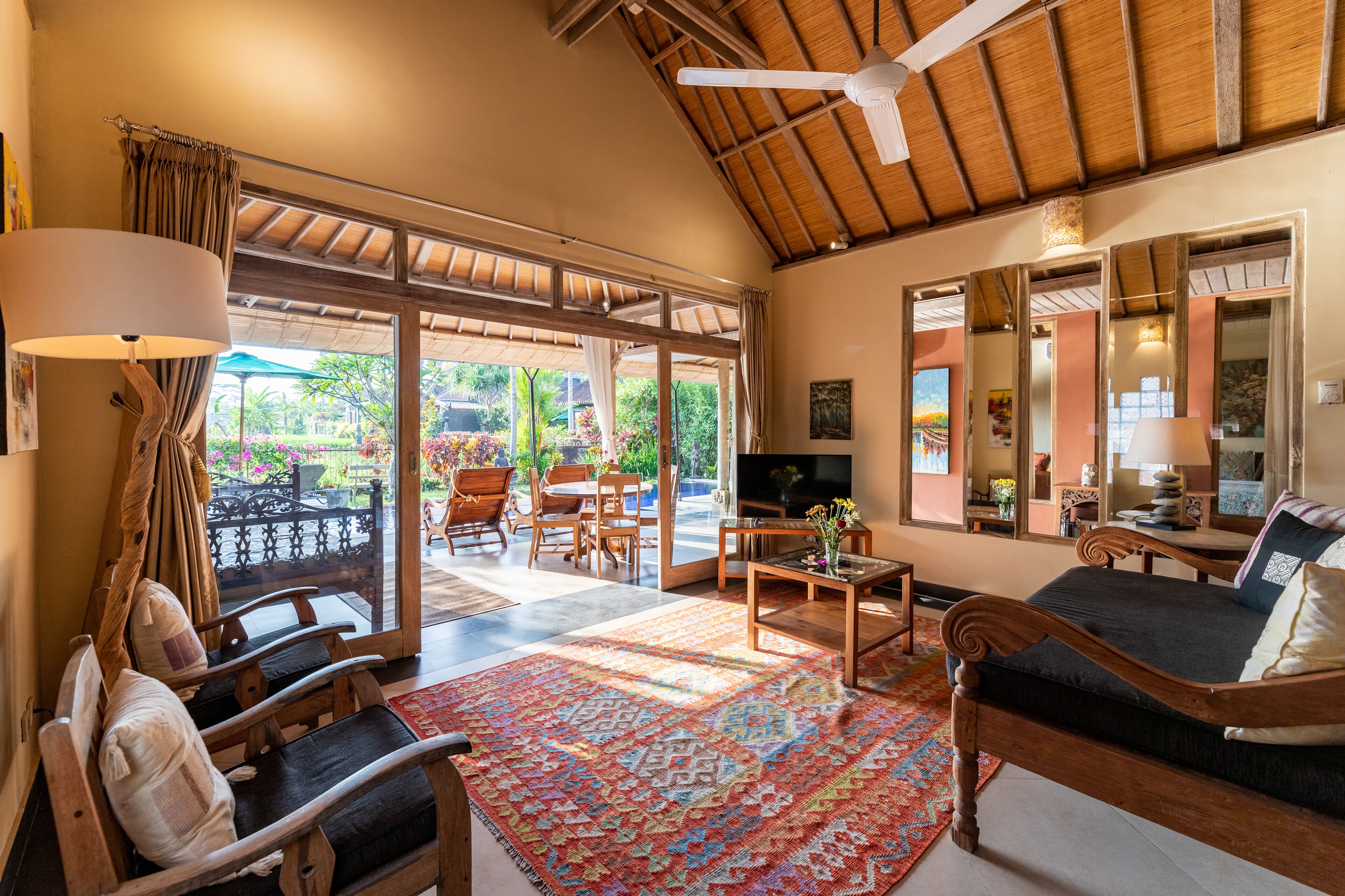 A spacious living area with a large sofa, a coffee table, and a TV. There is a balcony with a view of greenery and a dining area with chairs and a table.