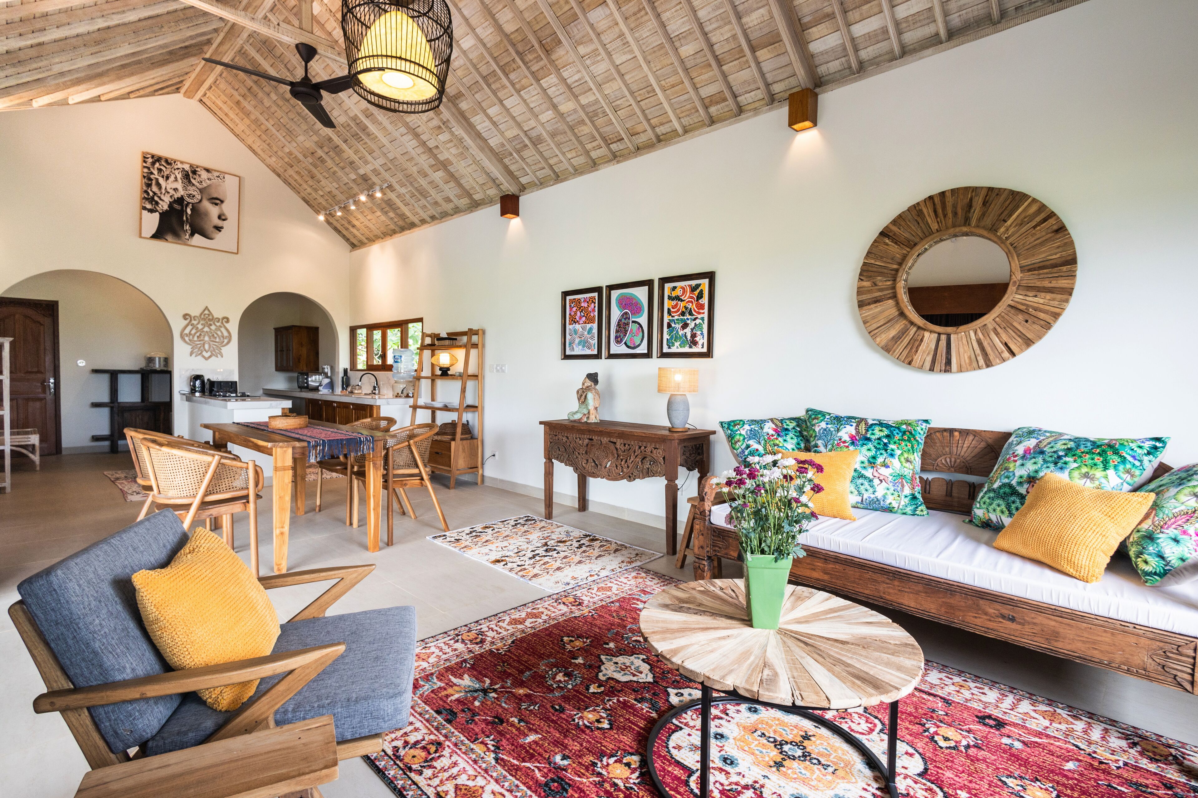 A spacious living area with a wooden ceiling, a round mirror, a sofa with colorful cushions, a dining table with chairs, and a kitchen in the background.