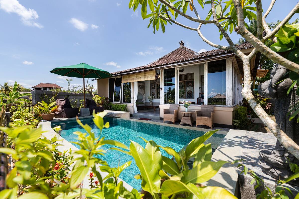Uma área à beira da piscina com um espaço de estar coberto, piscina e vegetação exuberante.