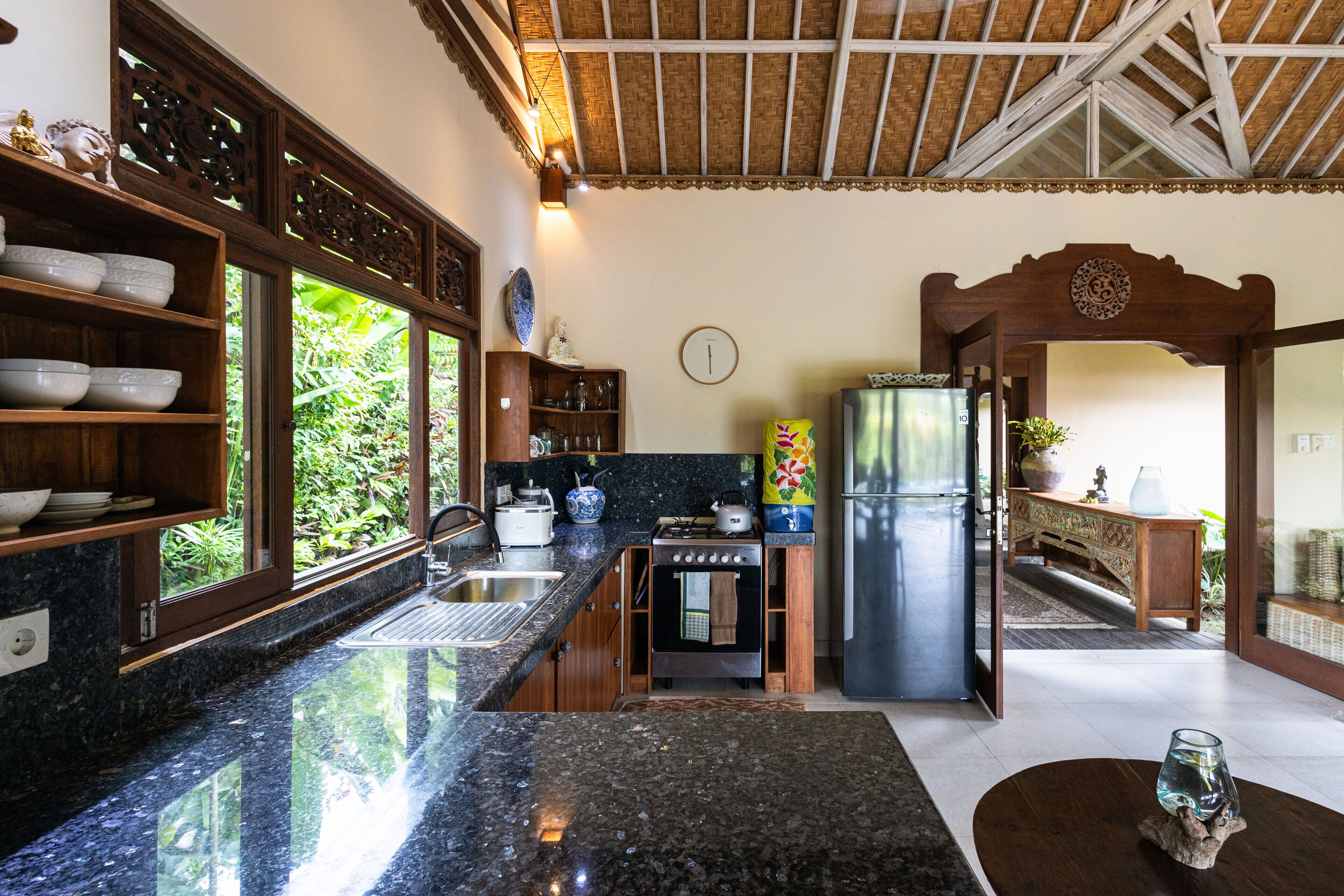 A kitchen with granite countertops, wooden cabinets, a stainless steel refrigerator, and a window with a view of greenery.
