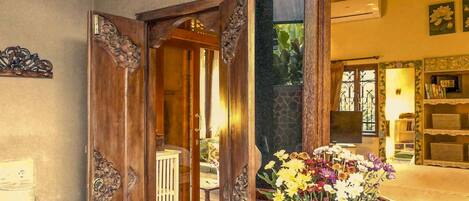 Una porta in legno con decorazioni elaborate, un soggiorno con un divano e un tavolino, e un vaso verde con fiori.