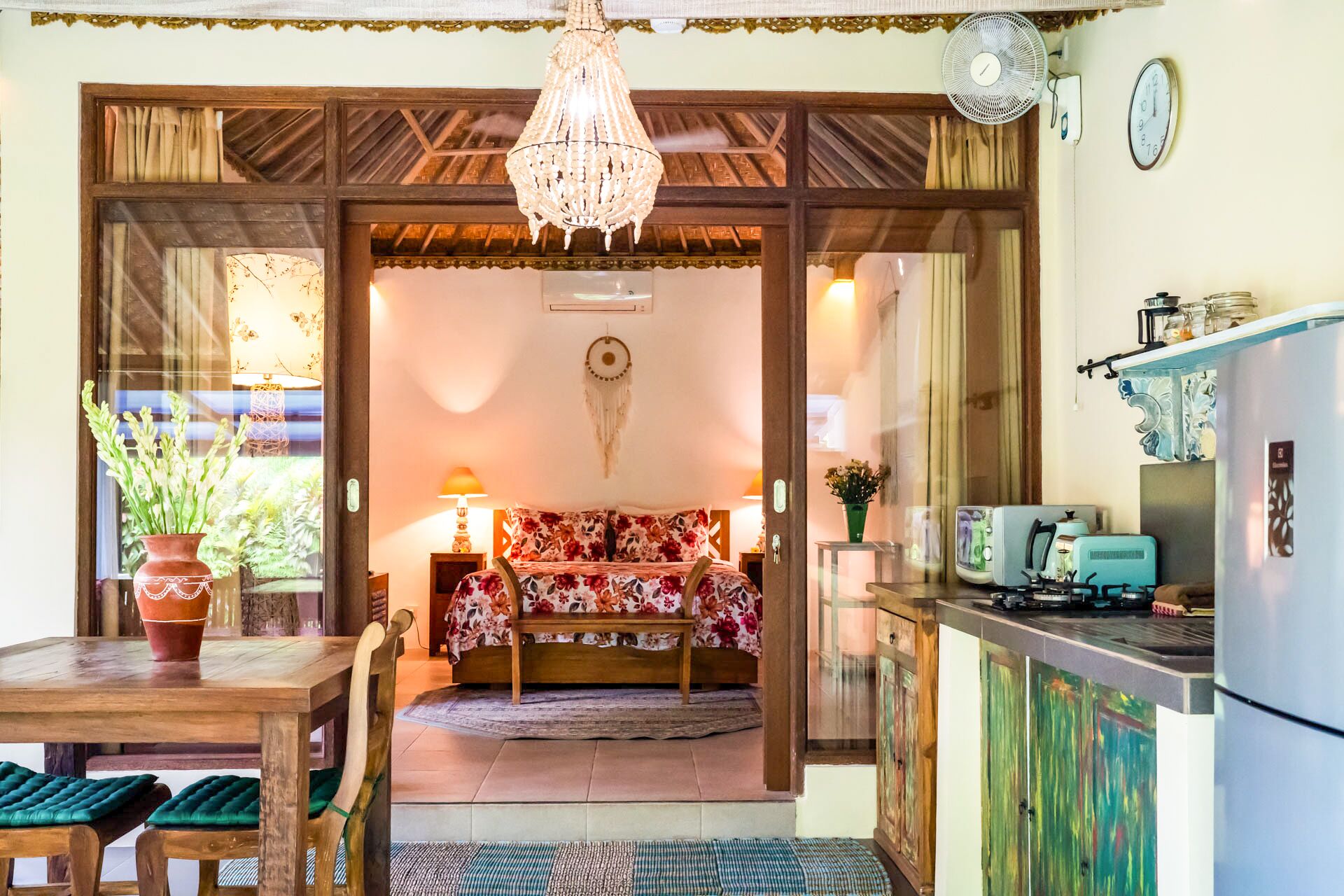 A cozy room with a bed, a wooden dining table with chairs, a chandelier, a kitchen area with a refrigerator, and a clock on the wall.