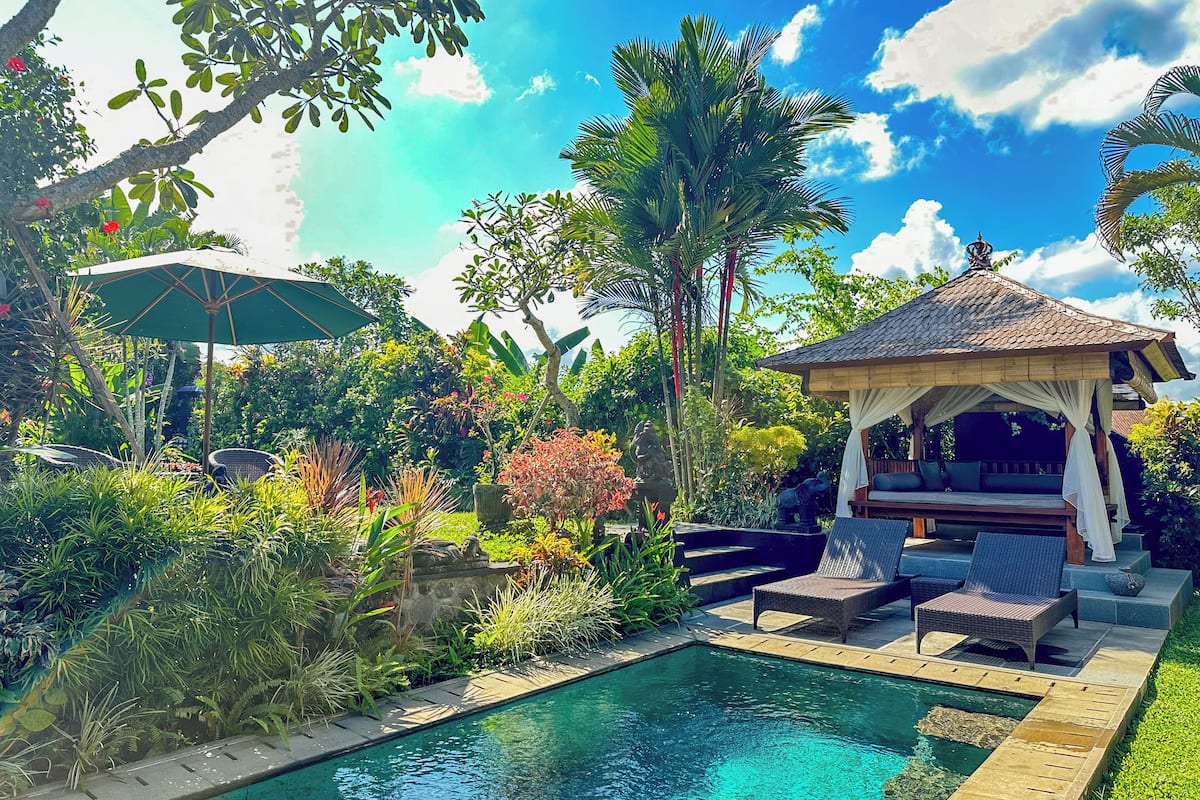 Uma área à beira da piscina com espreguiçadeiras, um gazebo e vegetação exuberante.