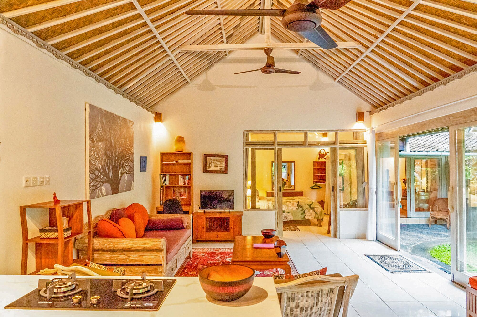 A cozy living room with a high ceiling, a ceiling fan, a fireplace, and a large window overlooking a garden.