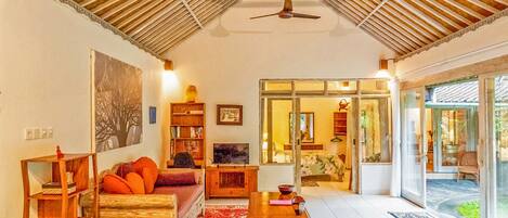 A cozy living room with a high ceiling, a ceiling fan, a fireplace, and a large window overlooking a garden.