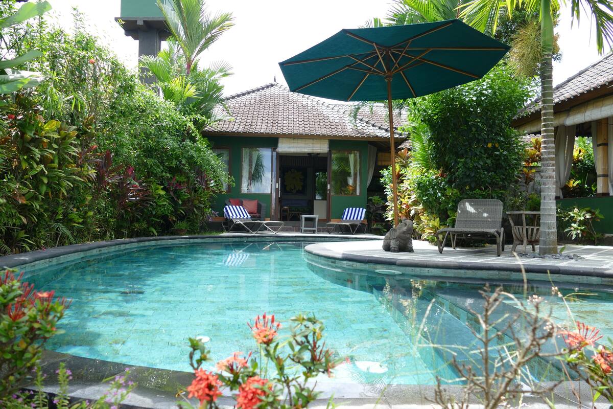Piscina com espreguiçadeiras e guarda-sol, cercada por vegetação exuberante e um edifício com telhado telhado.