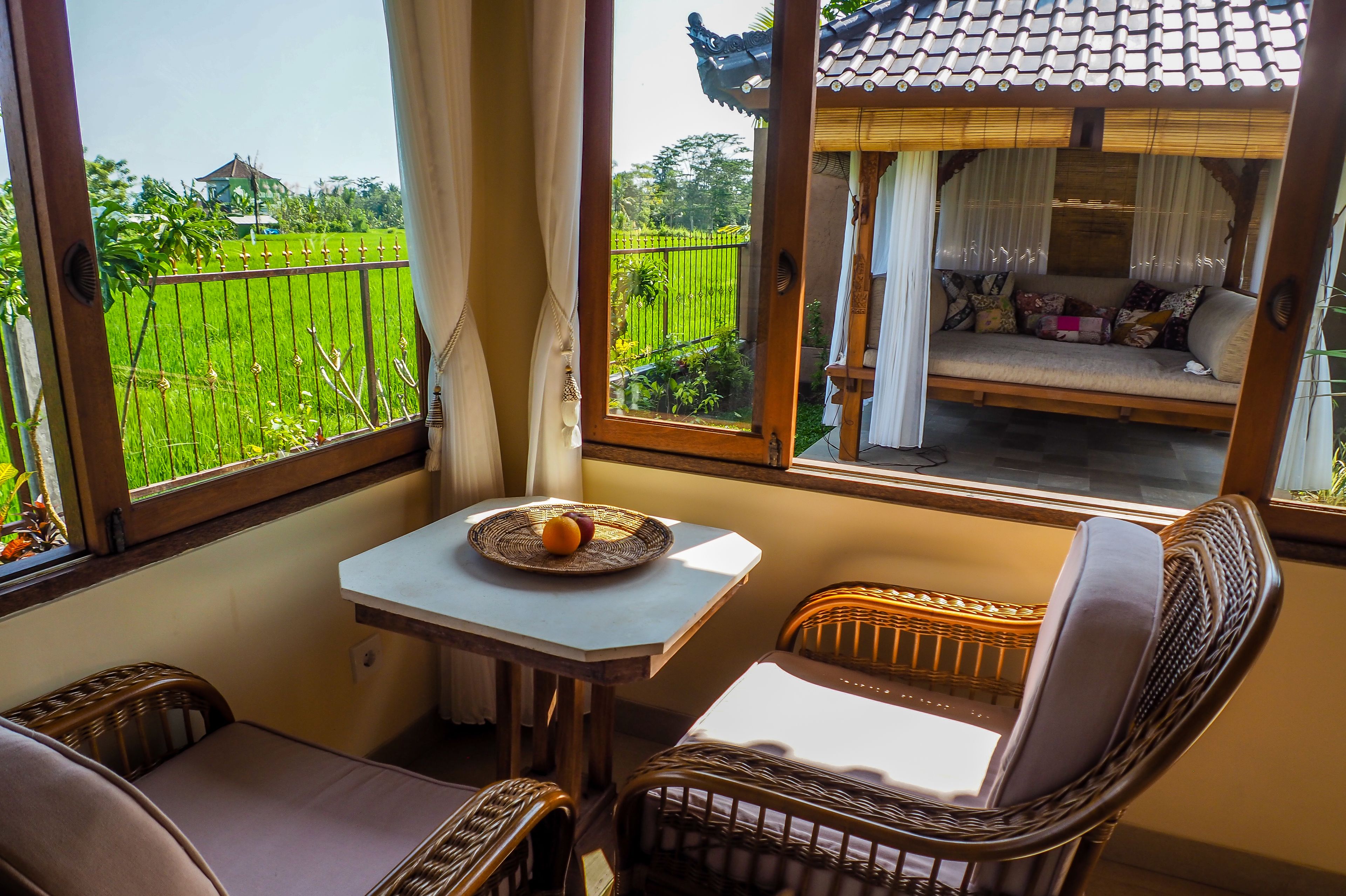 A room with a view of a green field, a small table with a fruit bowl, wicker furniture, and a traditional-style window with a bamboo structure.