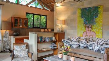 A cozy living room with a wooden sofa, a rattan chair, and a bookshelf filled with books. A large painting adorns the wall, and a ceiling fan is visible.