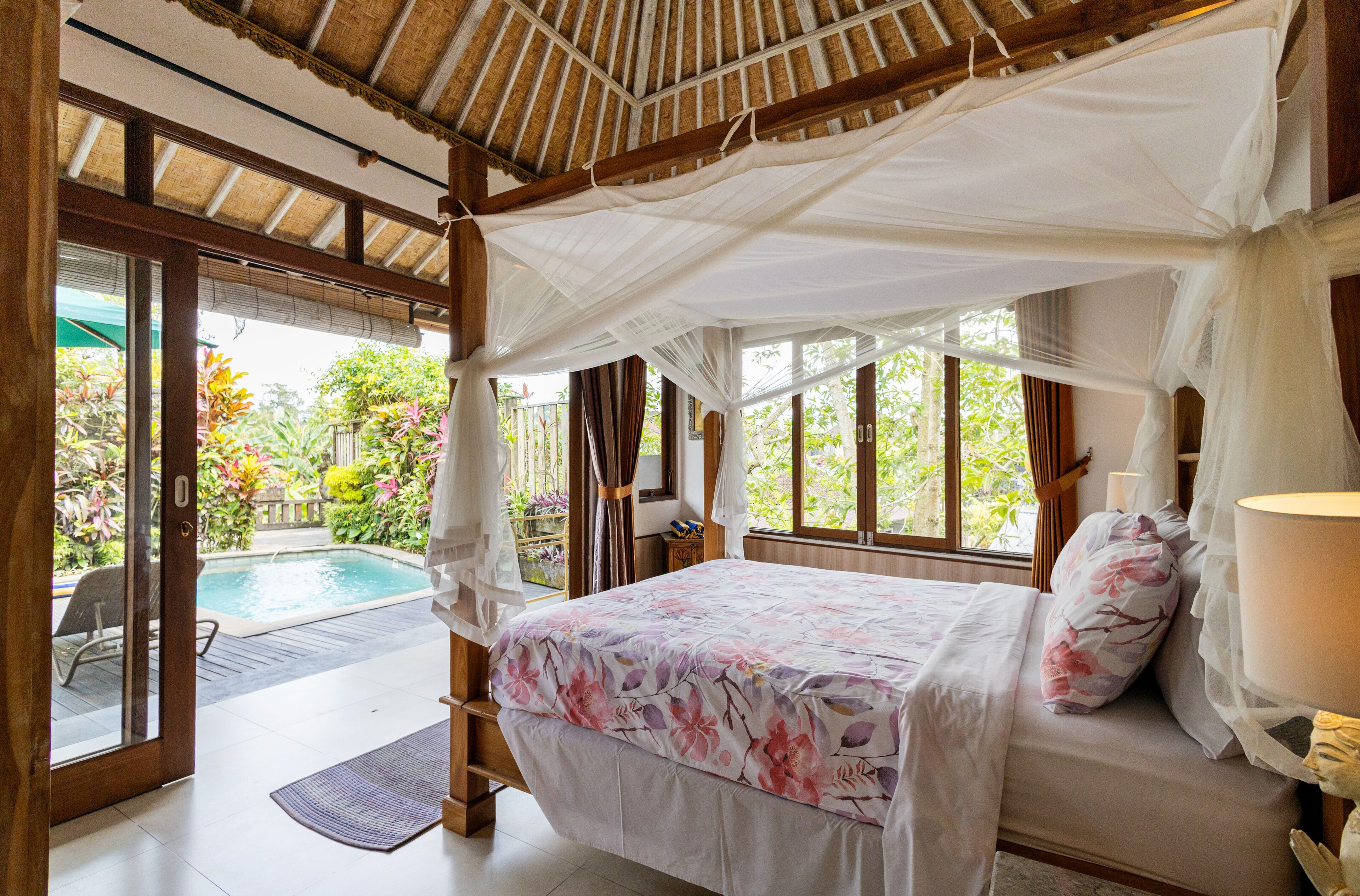 A bedroom with a bed, a bedside table, a lamp, a window, and a view of an outdoor swimming pool.