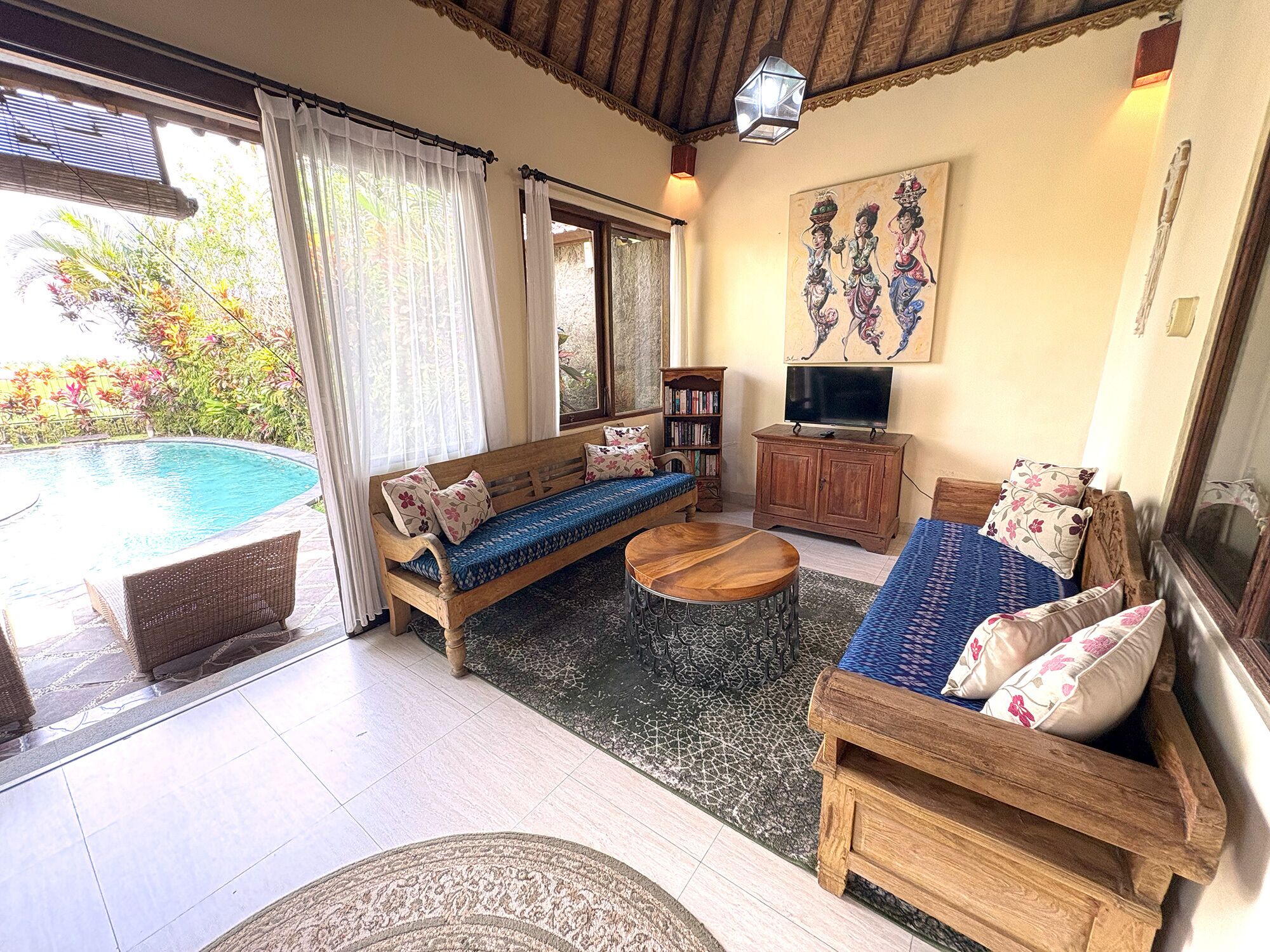 A living room with a sofa, a coffee table, and a TV stand. There is a pool visible through the glass doors.