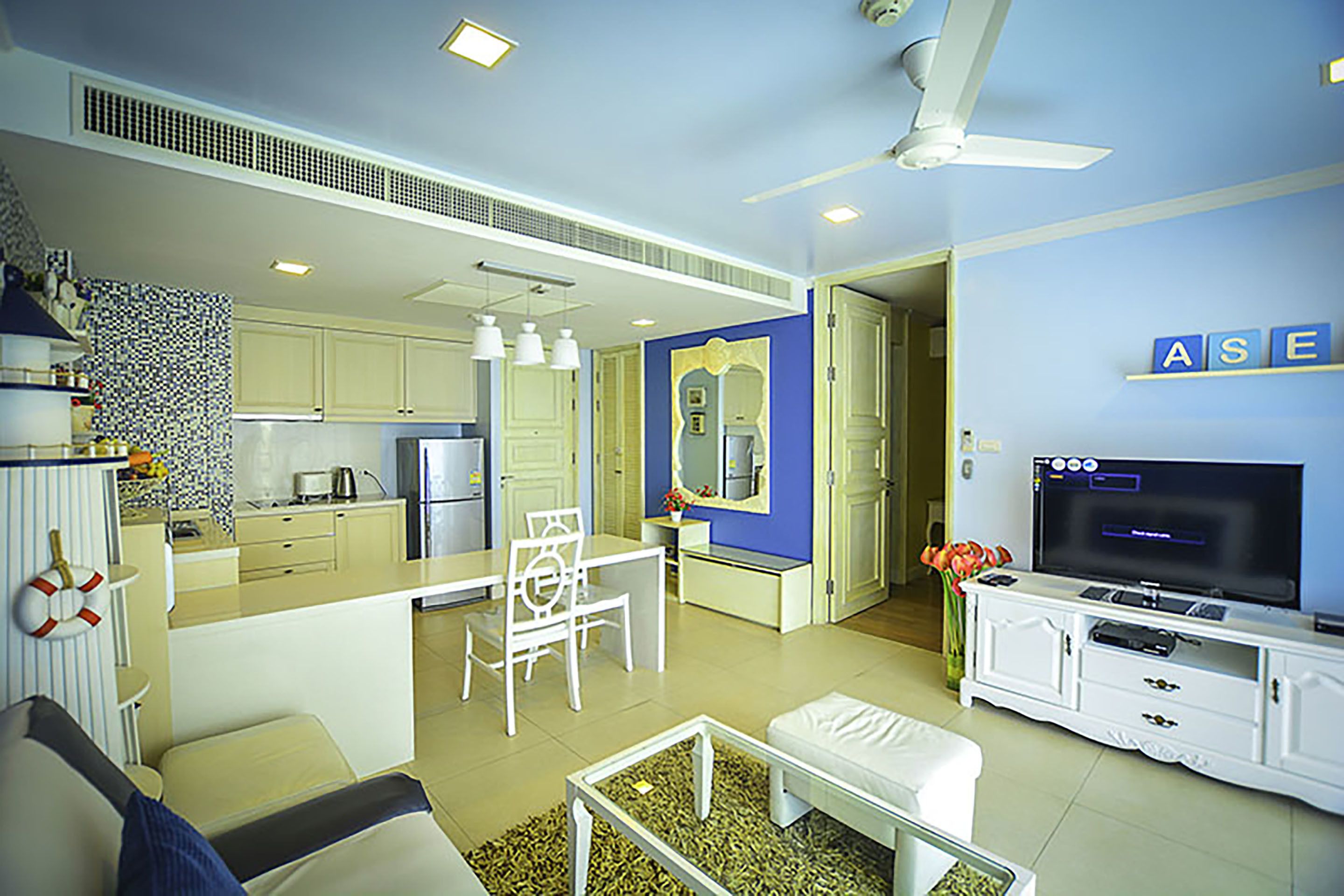 Una sala de estar moderna con un sofá, una mesa de centro y un televisor sobre un mueble bajo. Hay una zona de comedor con una mesa y sillas, y una cocina al fondo.