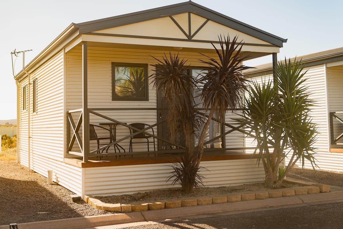 Une maison mobile de plain-pied dotée d’un balcon, de deux fenêtres et d’un petit espace patio aménagé avec des chaises et une table.