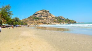 A beach with a rocky hill in the background, clear skies, and people walking along the shore. - The Sanctuary Hua Hin by Puppap (Hua Hin)