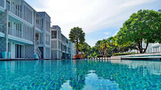 A swimming pool with clear blue water, surrounded by multi-story buildings with balconies and a tree-lined pathway. - Malibu Hua Hin by Puppap (Hua Hin)