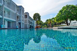 A swimming pool with clear blue water, surrounded by multi-story buildings with balconies and a tree-lined pathway. - Malibu Hua Hin by Puppap (Hua Hin)