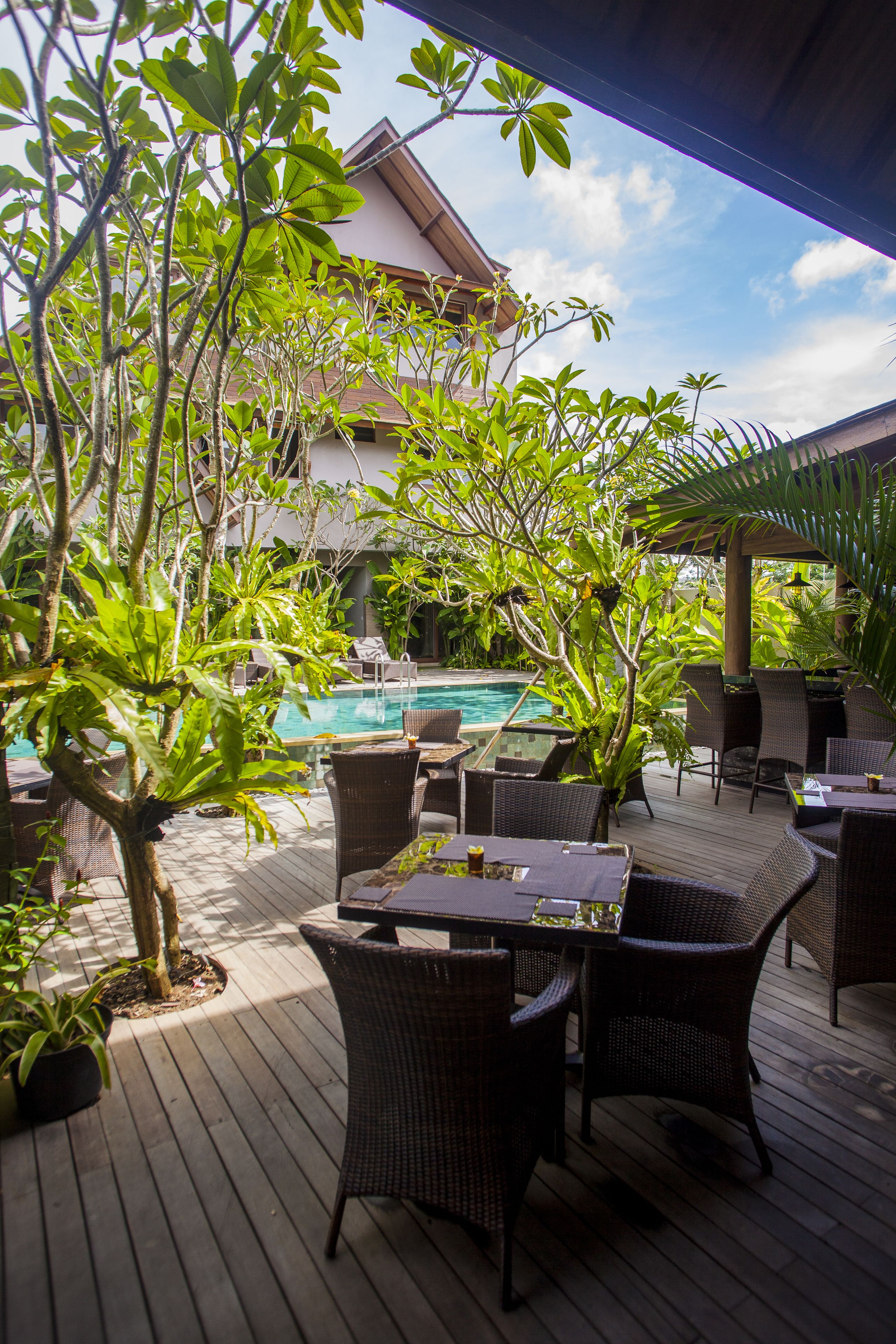 A wooden deck with wicker furniture, a pool, and lush greenery.