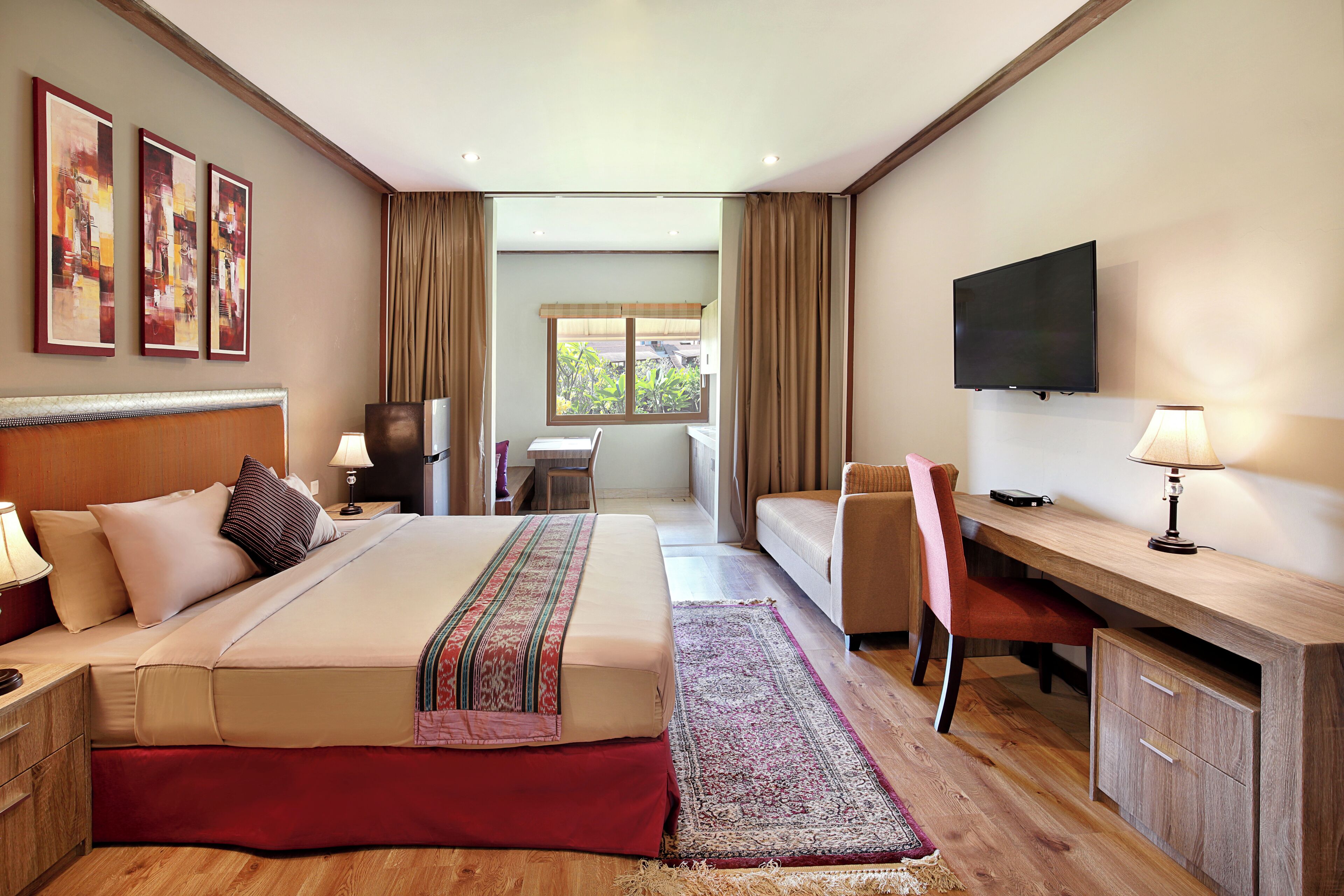 A hotel room with a bed, desk, chair, TV, and a view of greenery outside.
