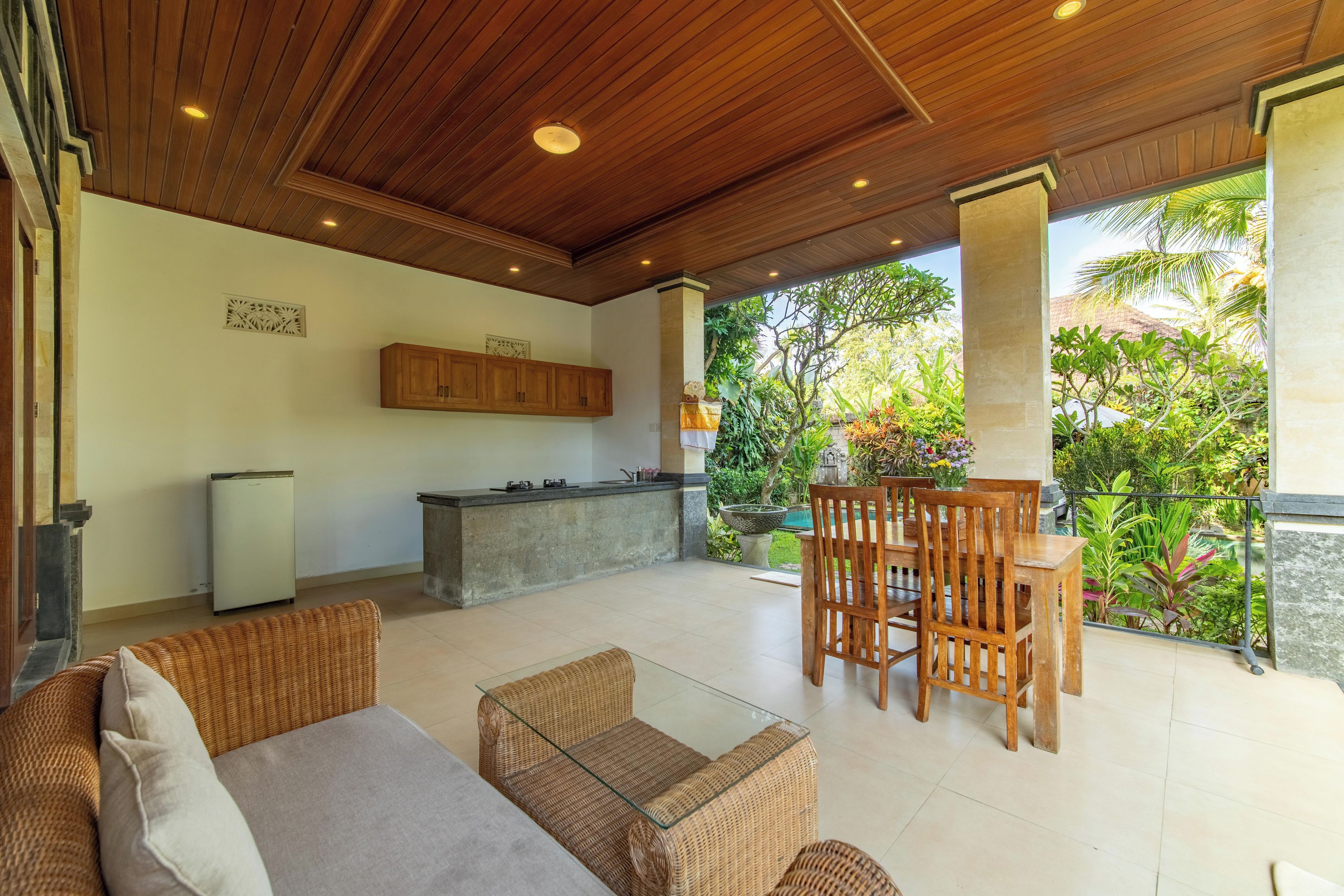 A spacious outdoor area with a wooden ceiling, a dining table with chairs, a kitchen area, and a view of lush greenery.