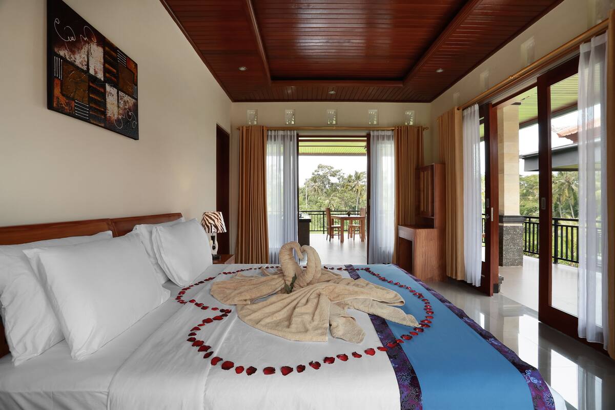 A hotel room with a bed, a painting on the wall, and a balcony with a view of trees.