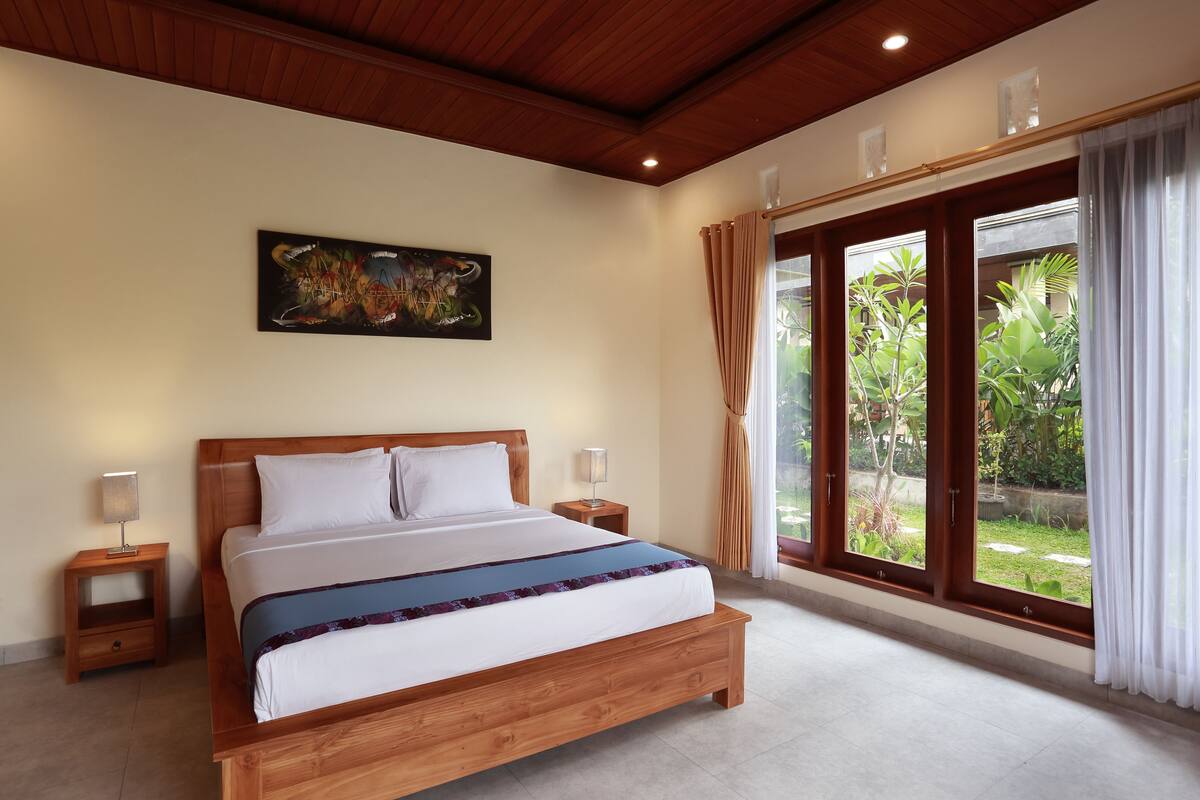 A bedroom with a wooden bed, a nightstand, a lamp, a painting on the wall, and a large window with curtains.
