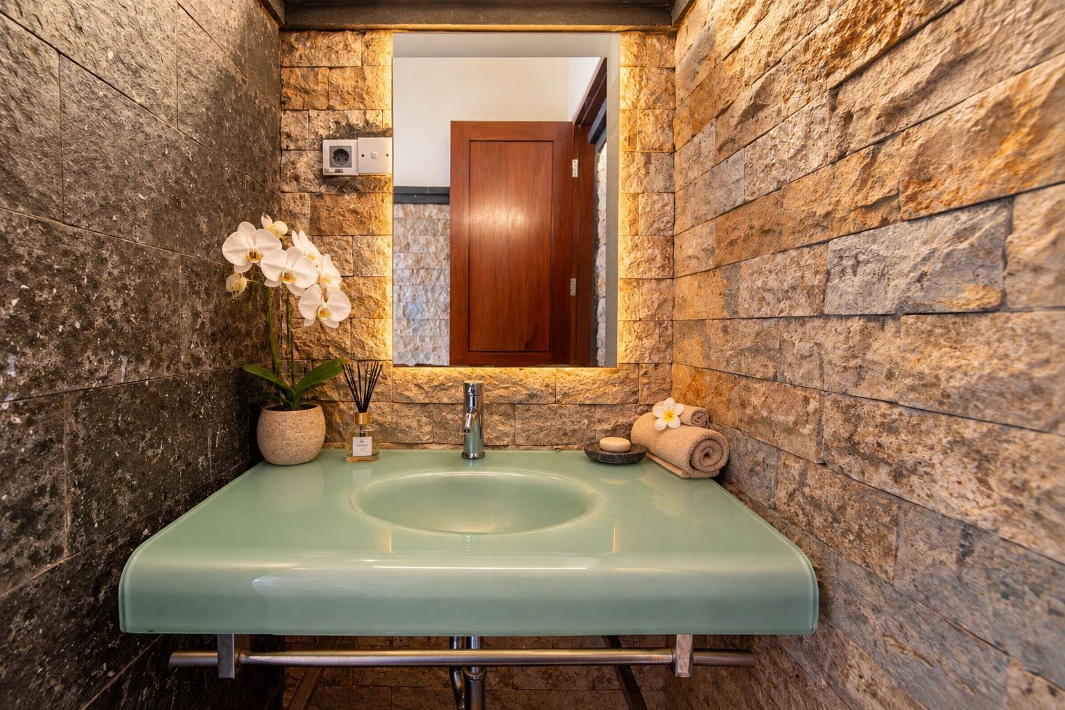 A bathroom with a stone wall, a green sink, a mirror, and a wooden door.