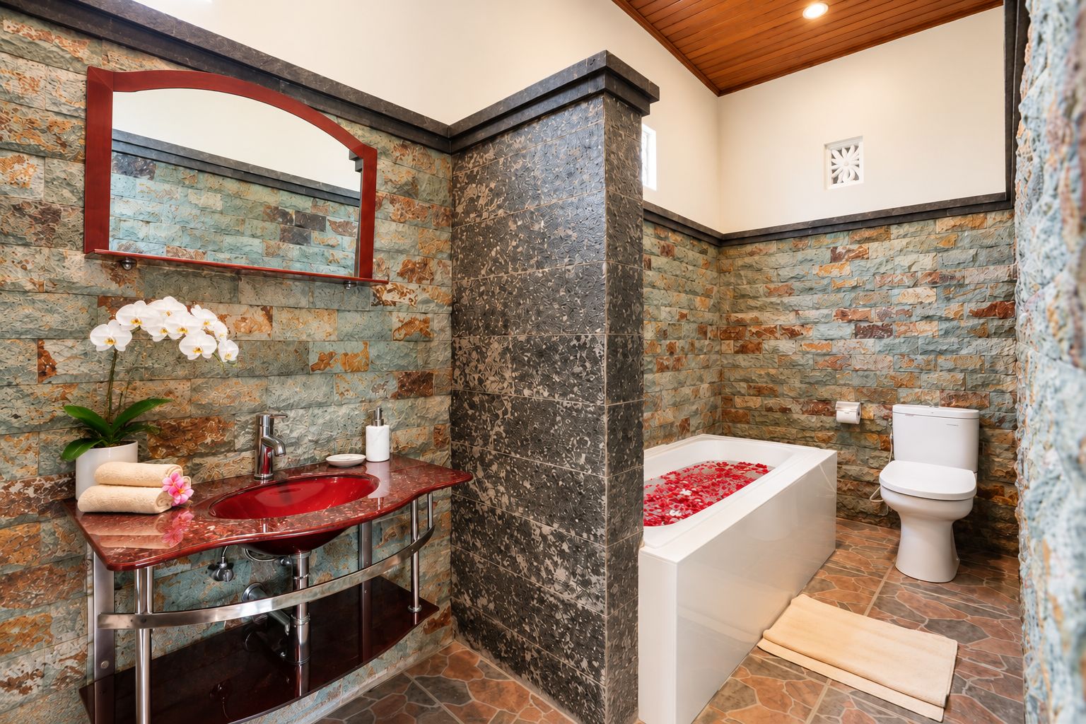 Ein Badezimmer mit Steinwand, Waschbecken in Rot, Toilette in Weiß und einer Badewanne mit Rosenblättern.