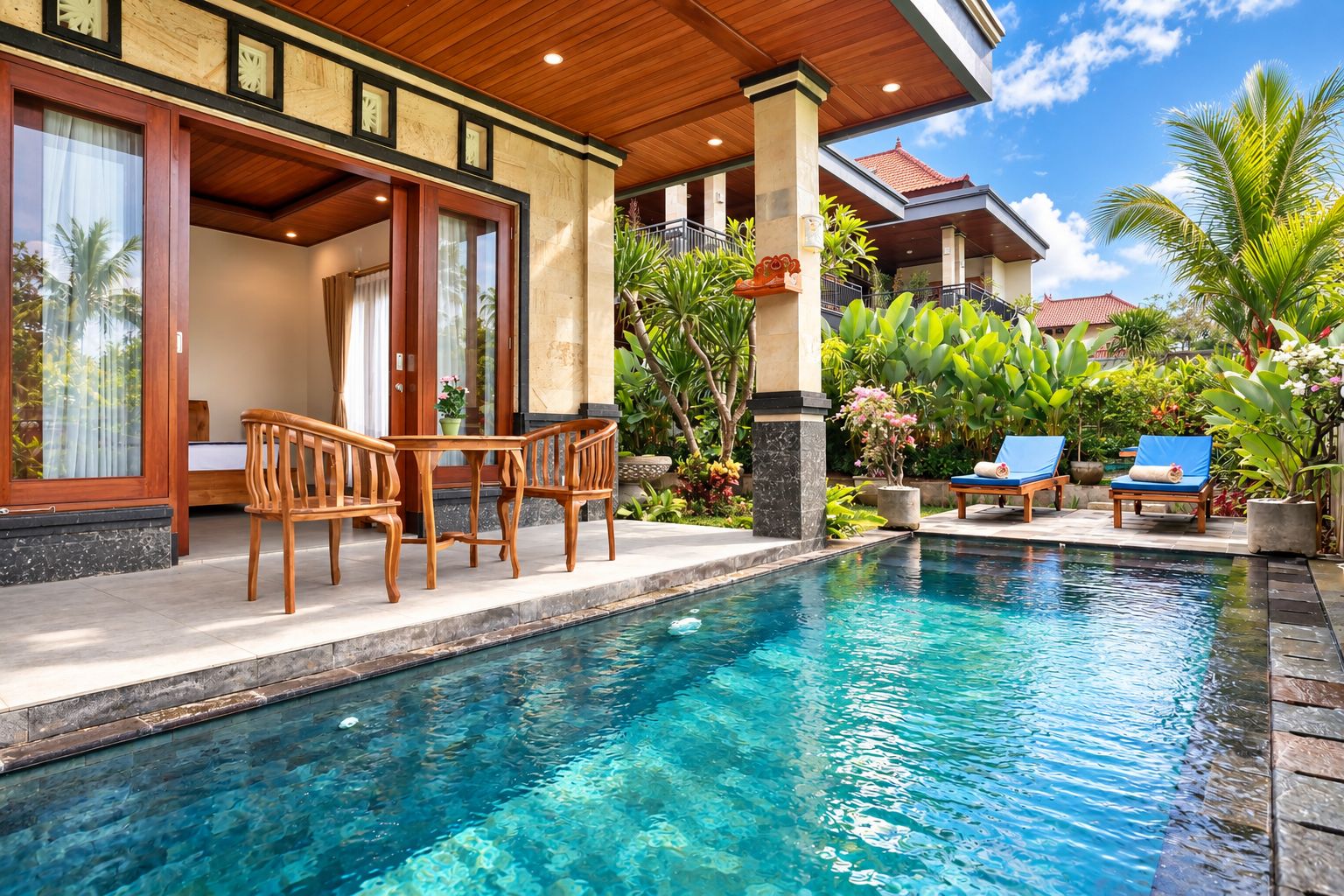 A swimming pool with wooden deck chairs and a table, surrounded by lush tropical plants.