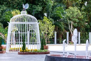Una jaula blanca con una estatua de pájaro en la parte superior, rodeada de plantas y una fuente de agua.