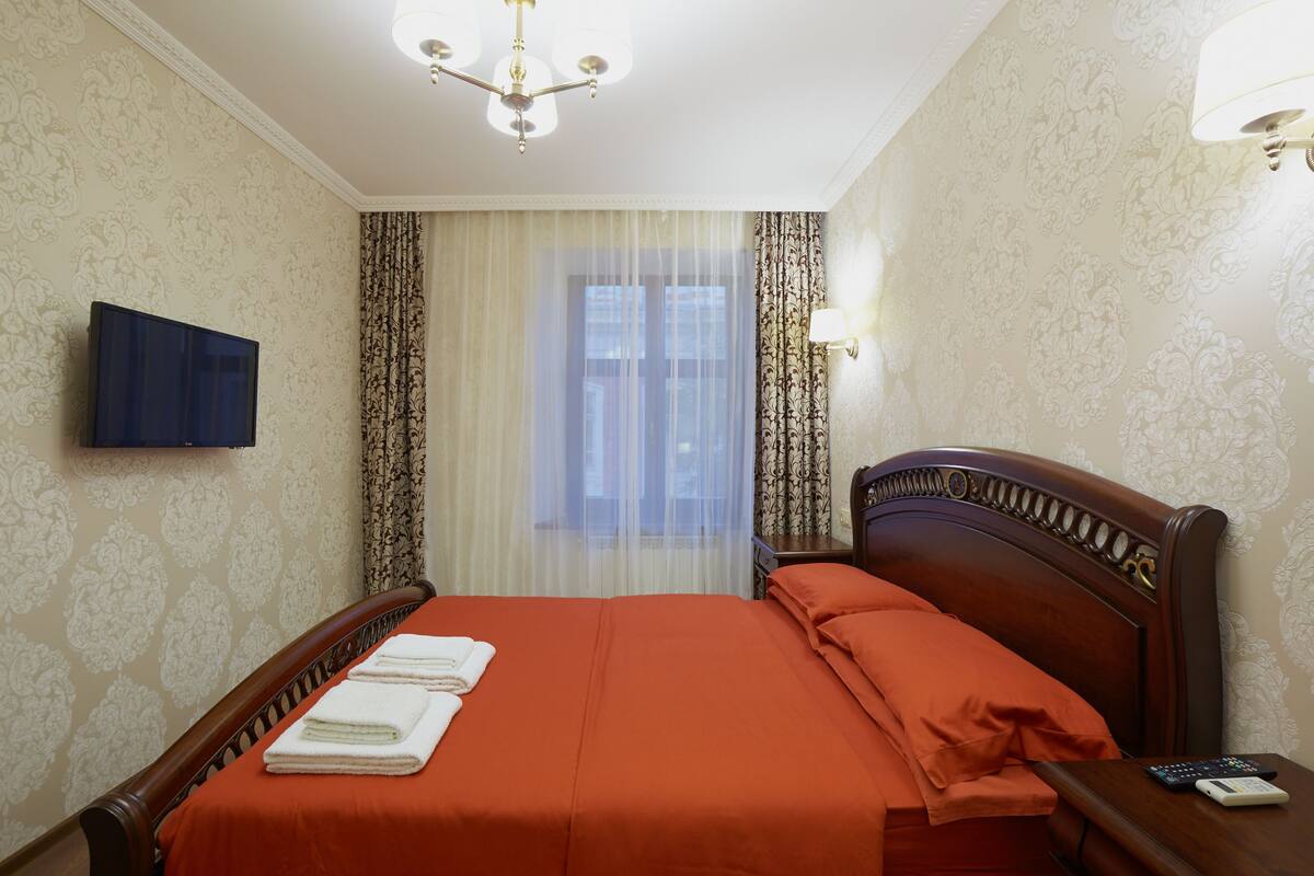 Quarto de hotel com cama, televisão fixada na parede, janela com cortinas e um candelabro.
