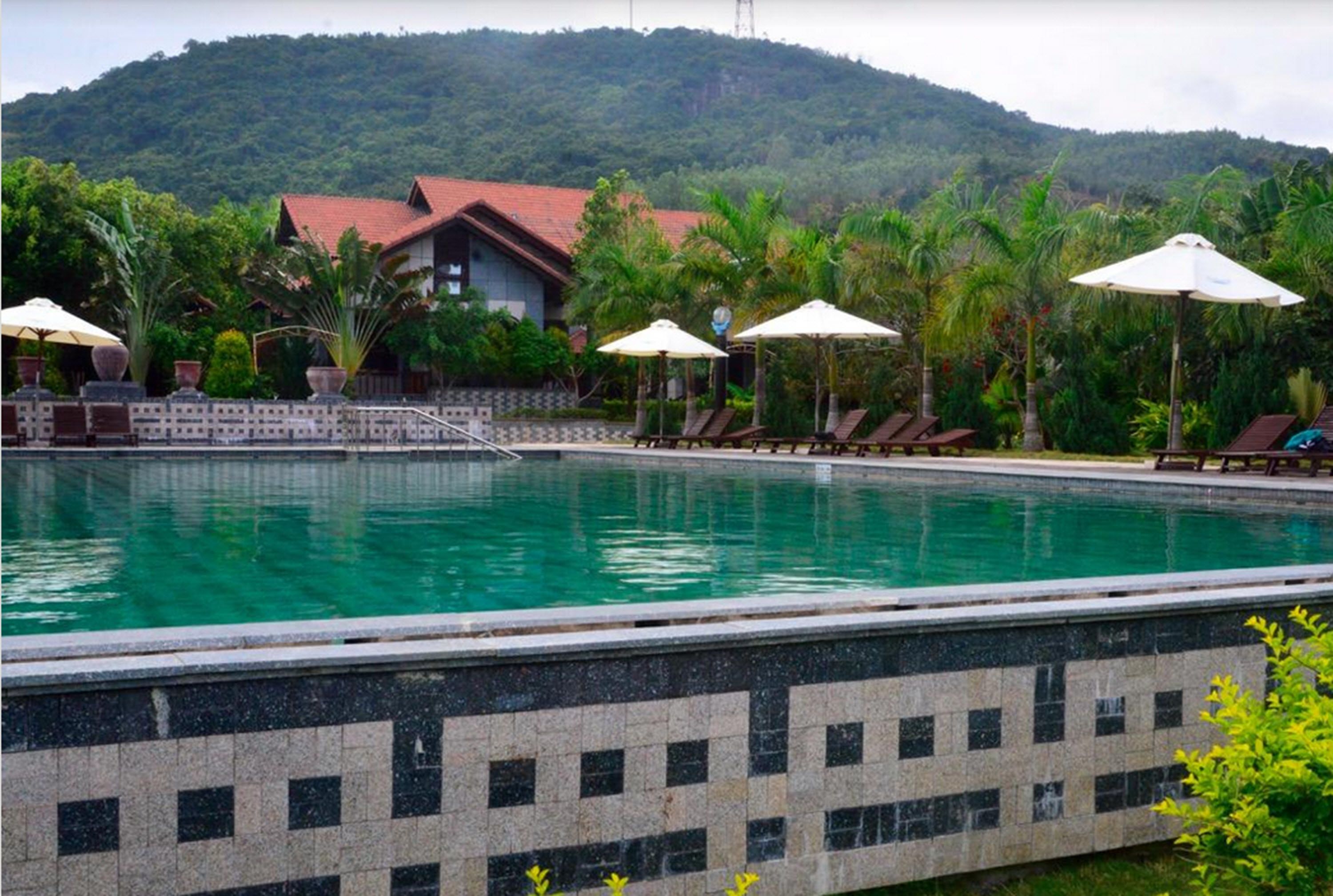Ein Swimmingpool mit Liegestühlen und Sonnenschirmen, umgeben von üppiger Vegetation und einer Hügelkulisse im Hintergrund.