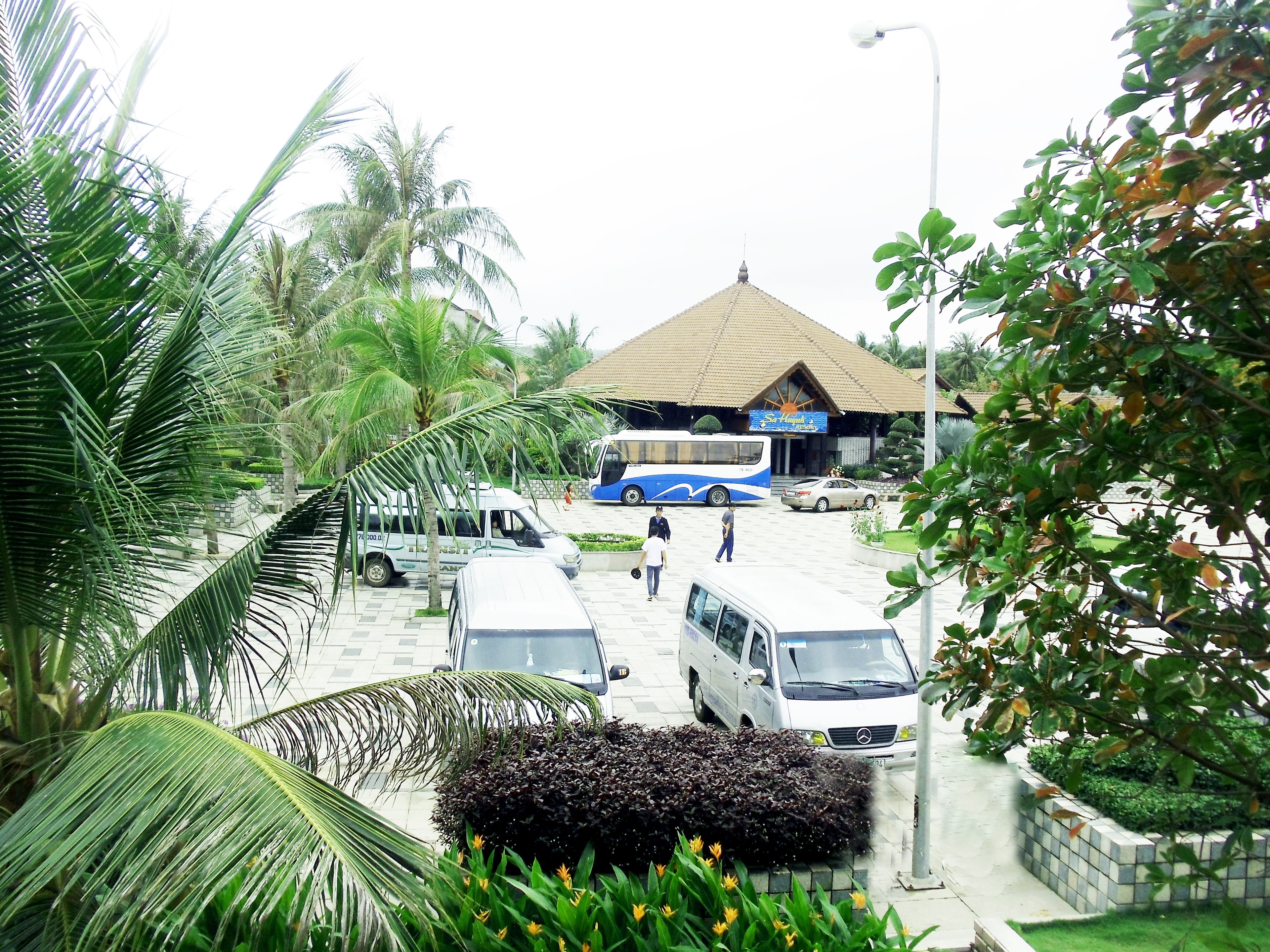 A bus station with a bus, a van, and a car parked. There are palm trees and a building with a tiled roof in the background.