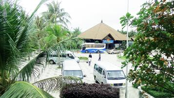 A bus station with a bus, a van, and a car parked. There are palm trees and a building with a tiled roof in the background.