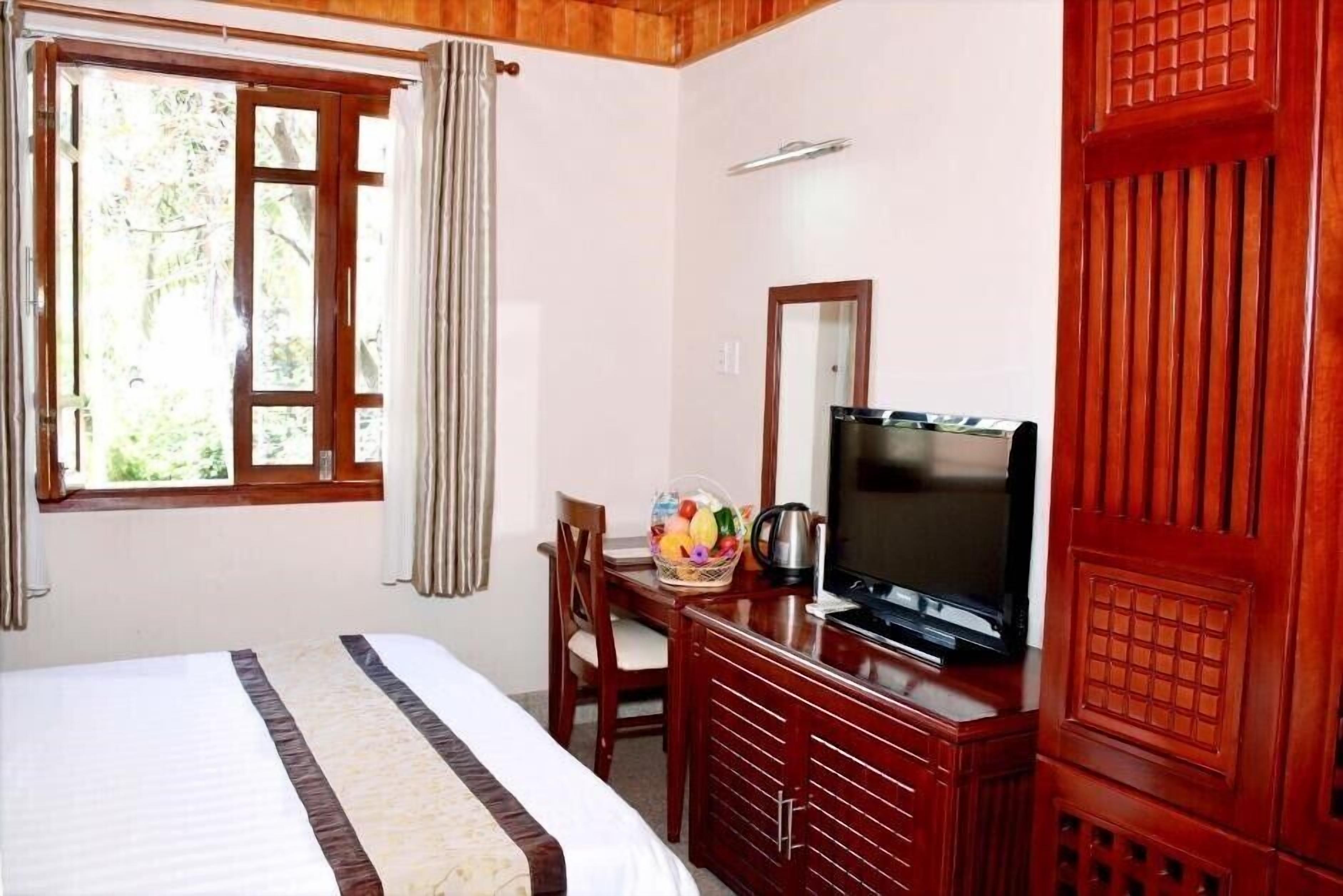 Habitación de hotel con cama, televisor, cómoda de madera, ventana con cortinas y vista a espacios verdes.