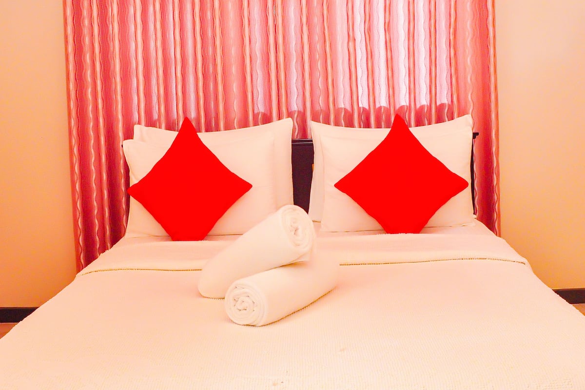 A hotel room with a bed, red pillows, and a rolled white towel.