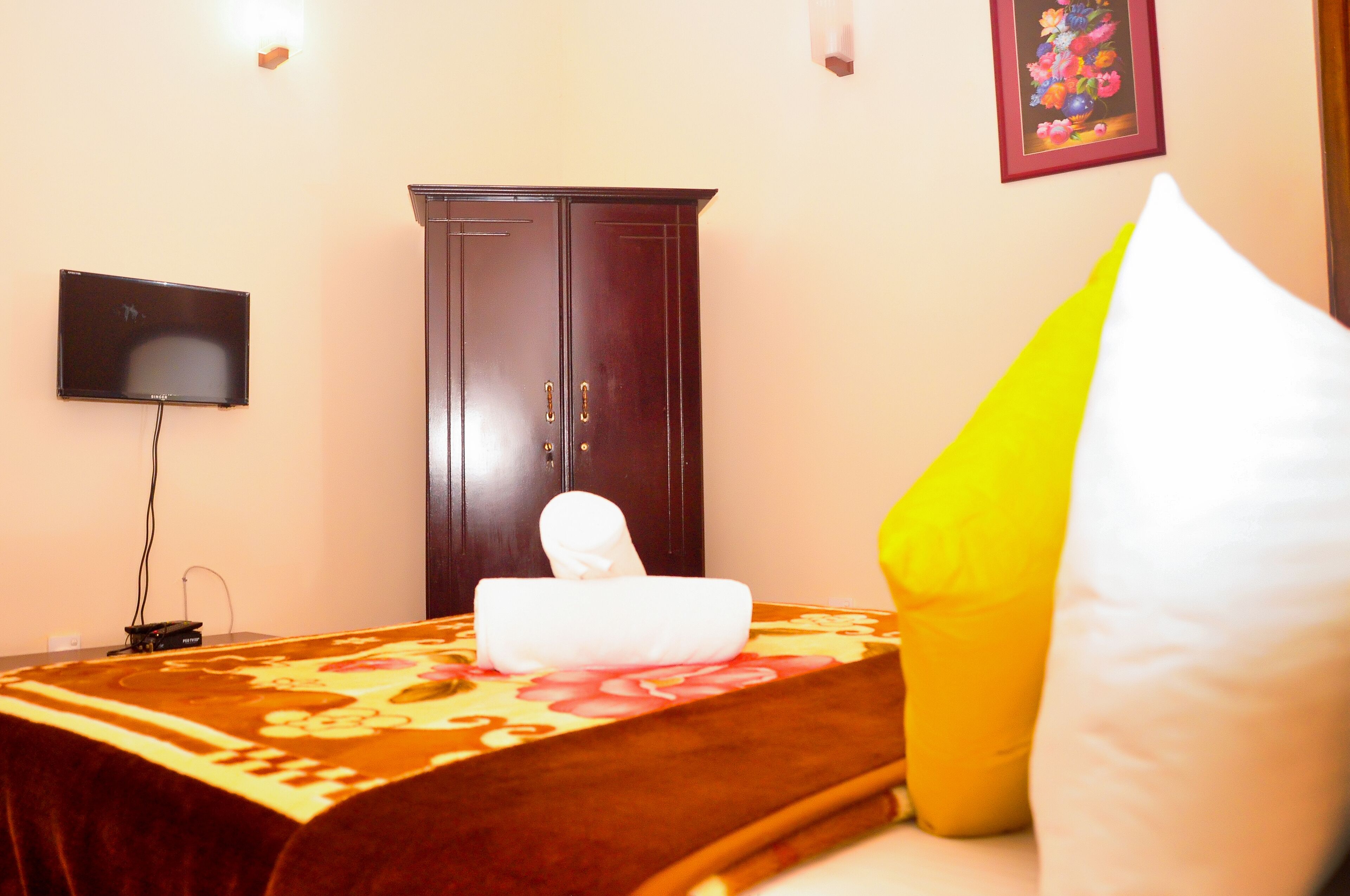 A hotel room with a bed, a flat-screen TV mounted on the wall, a wooden wardrobe, and a decorative pillow.