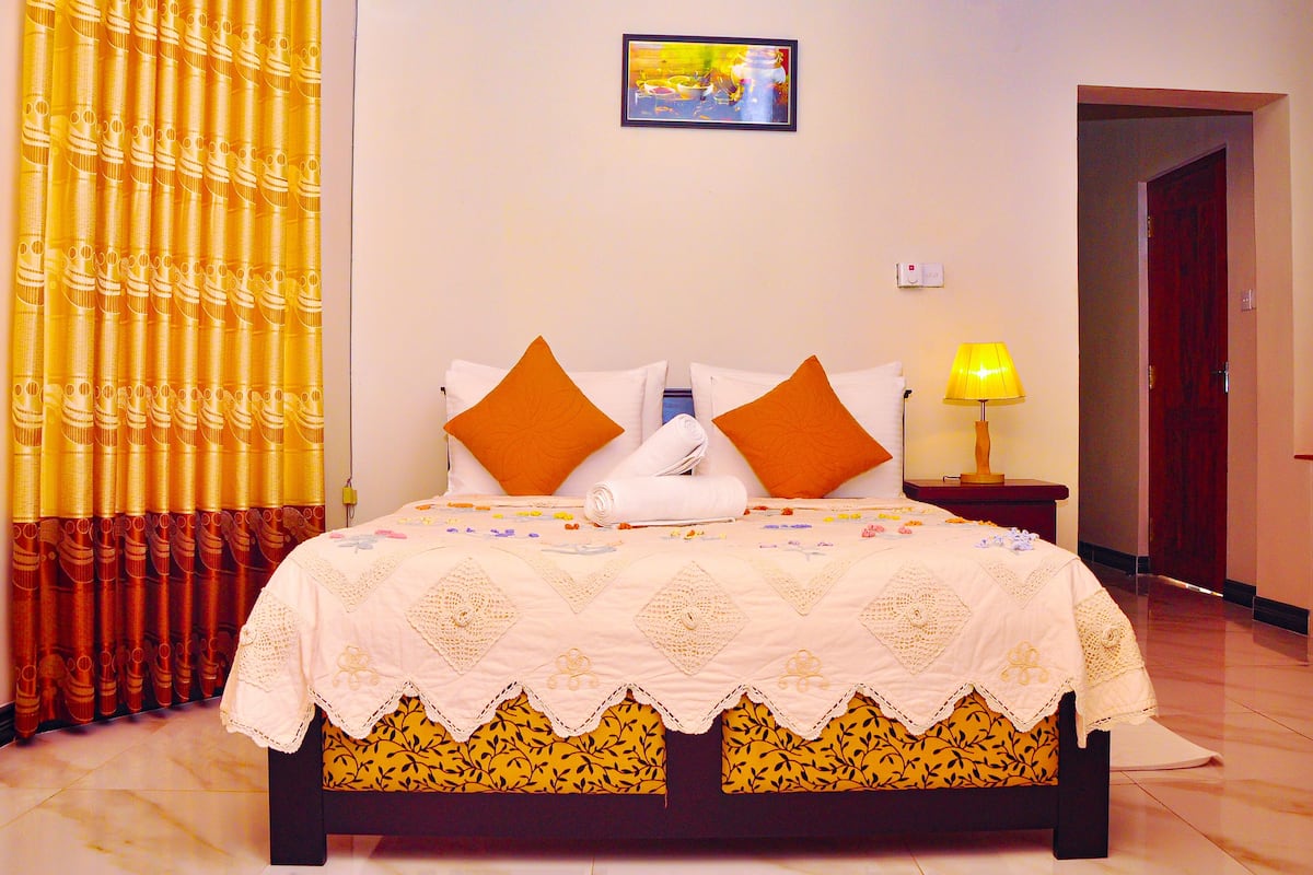 A neatly made bed with a white and yellow bedspread, two orange pillows, and a small bedside table with a lamp.