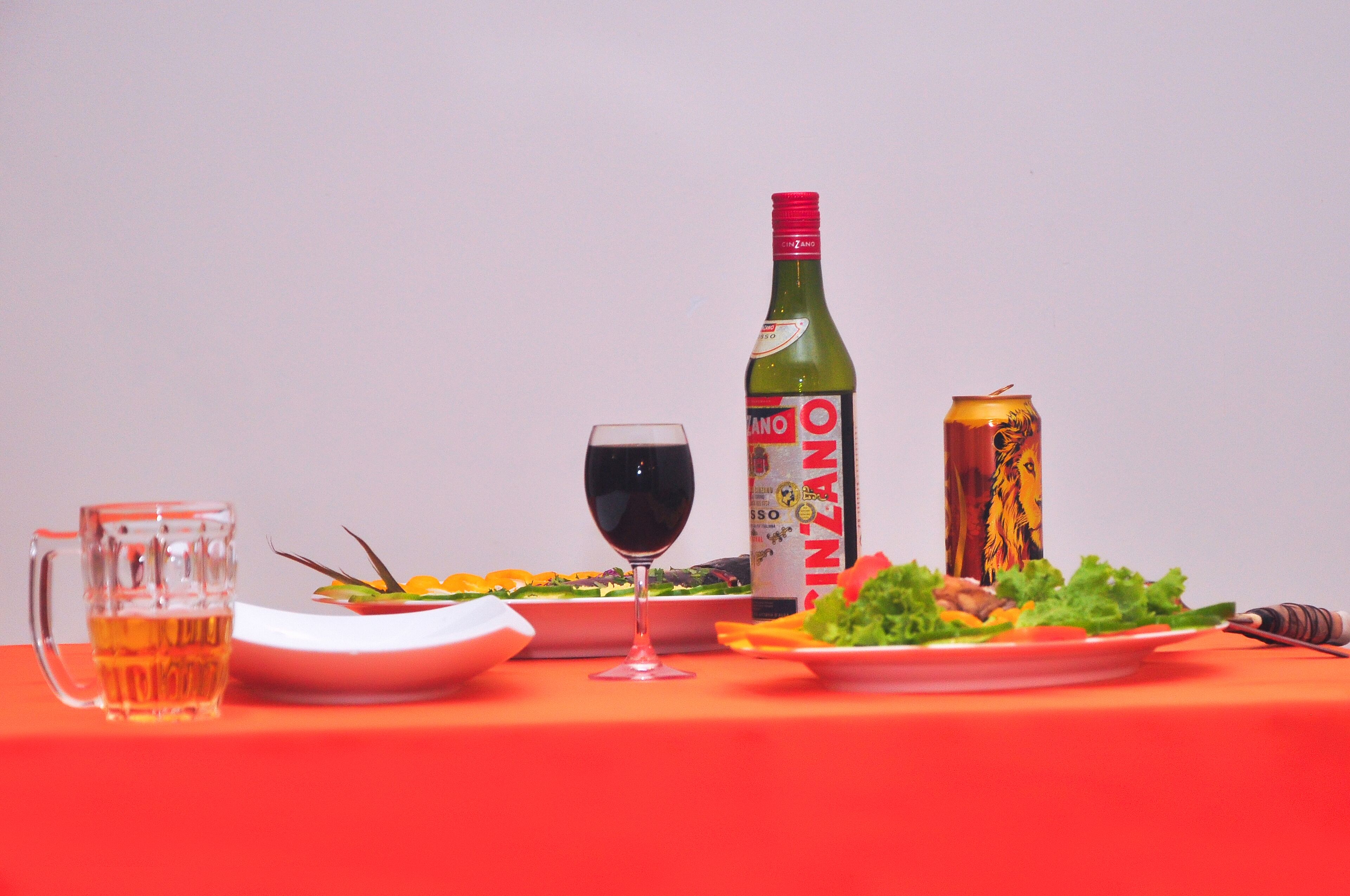 Una mesa con mantel rojo, una jarra de cerveza, una copa de vino, una botella de vino, una lata de cerveza y platos con comida.