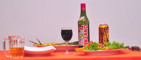 Una mesa con mantel rojo, una jarra de cerveza, una copa de vino, una botella de vino, una lata de cerveza y platos con comida.