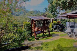 A rustic wooden swing chair in a lush garden with a metal roof structure and white plastic chairs. - New Kandy Residence (Kandy)