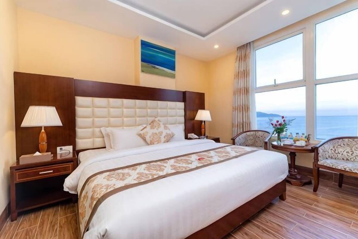Een hotelkamer met een groot bed, twee nachtkastjes, een stoel, een klein tafeltje met een vaas, en een raam met uitzicht op zee.