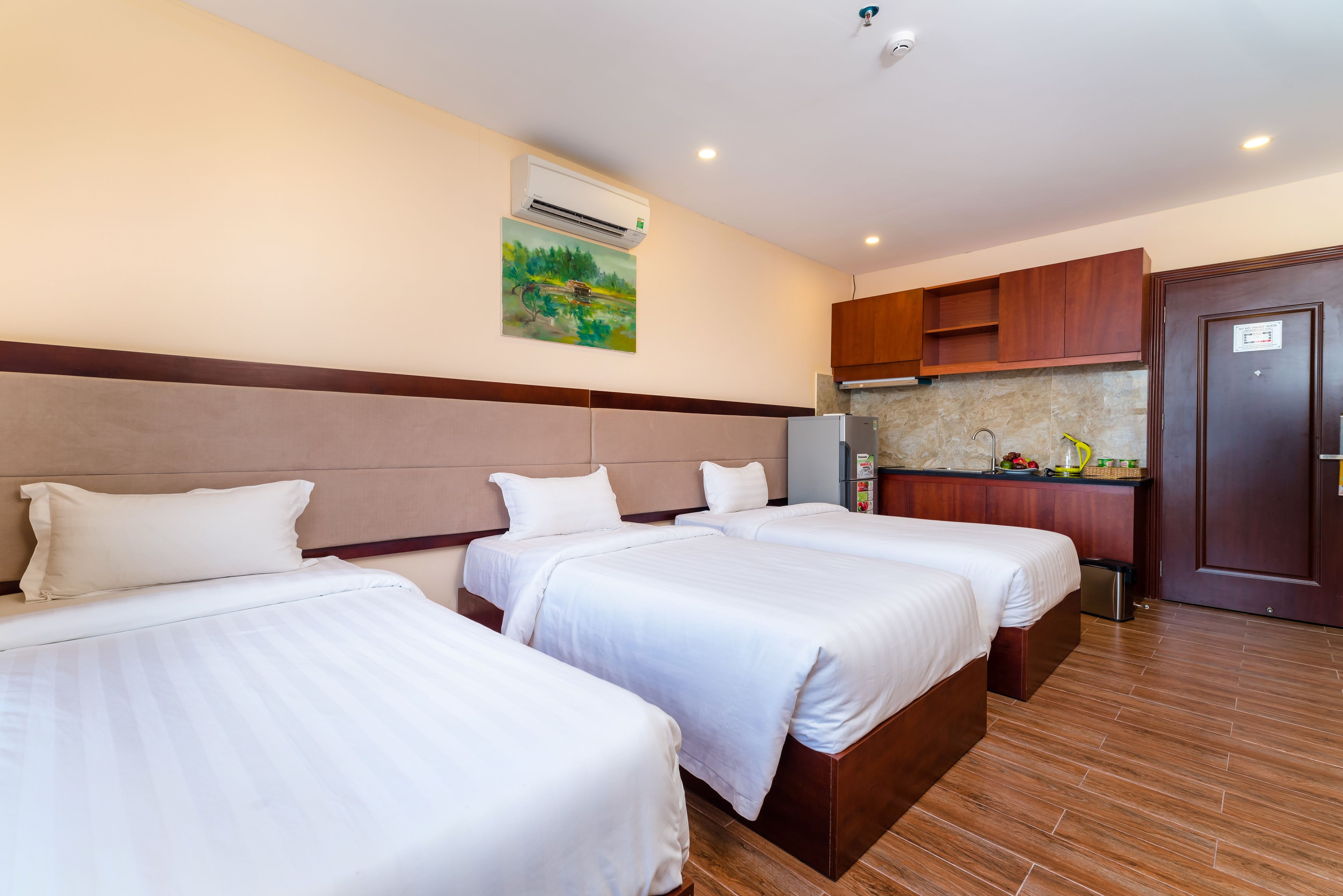 A hotel room with three beds, a kitchenette, and a painting on the wall.