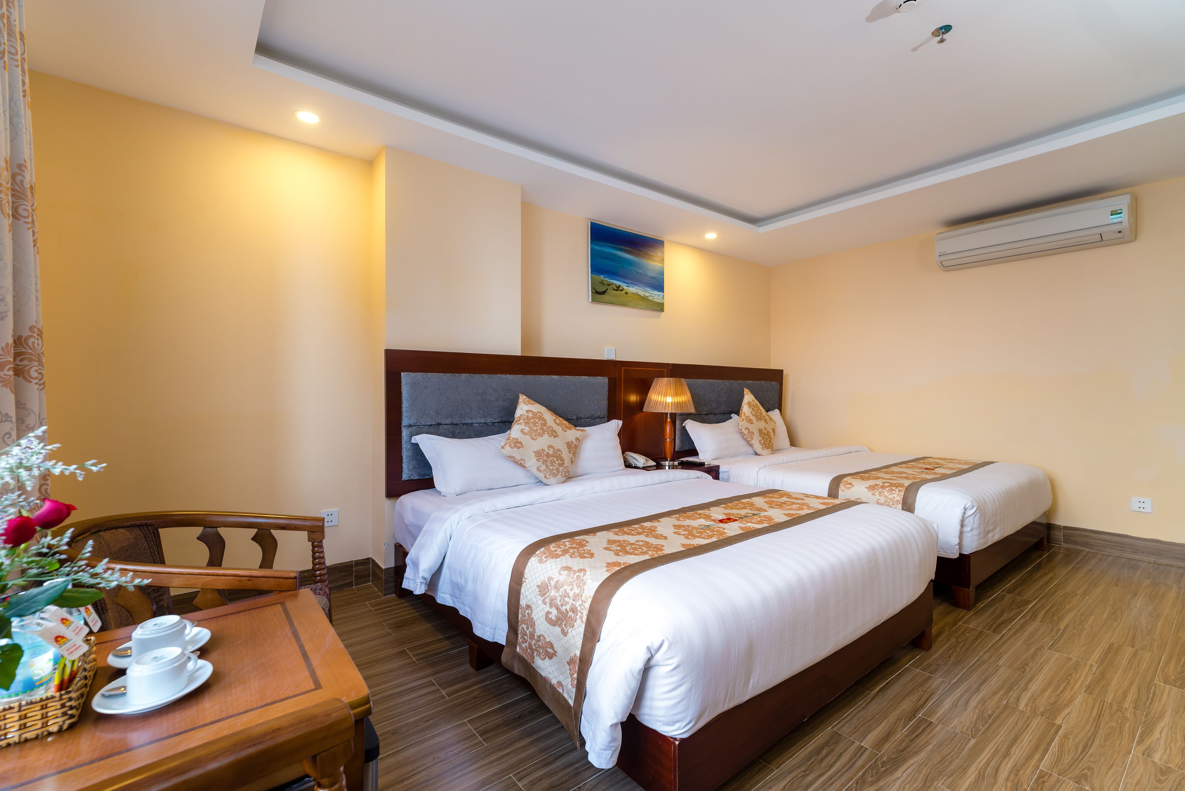 A hotel room with two beds, a wooden table with a basket of items, and a painting on the wall.
