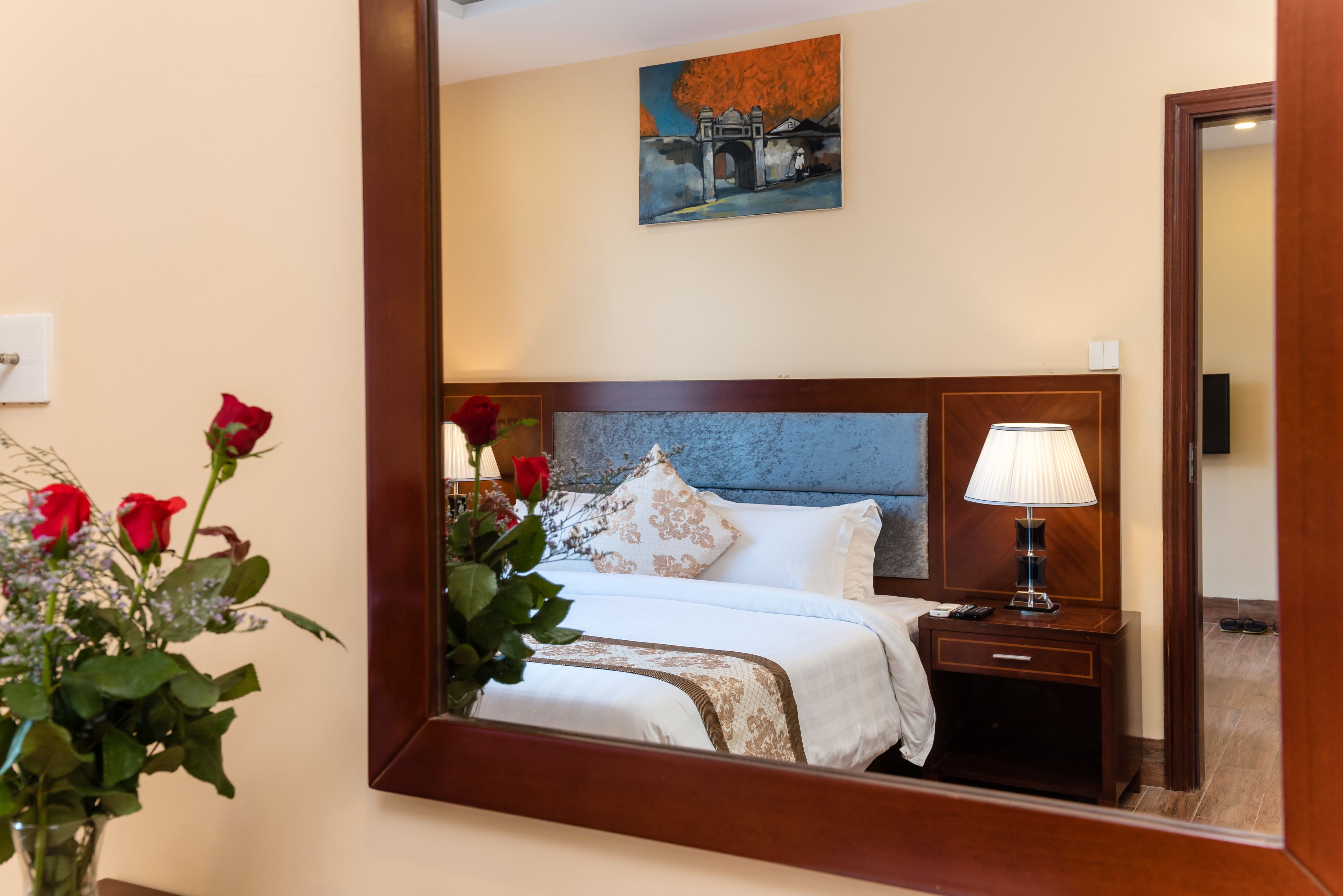 A hotel room with a bed, a nightstand, a lamp, a painting on the wall, and a vase with flowers.