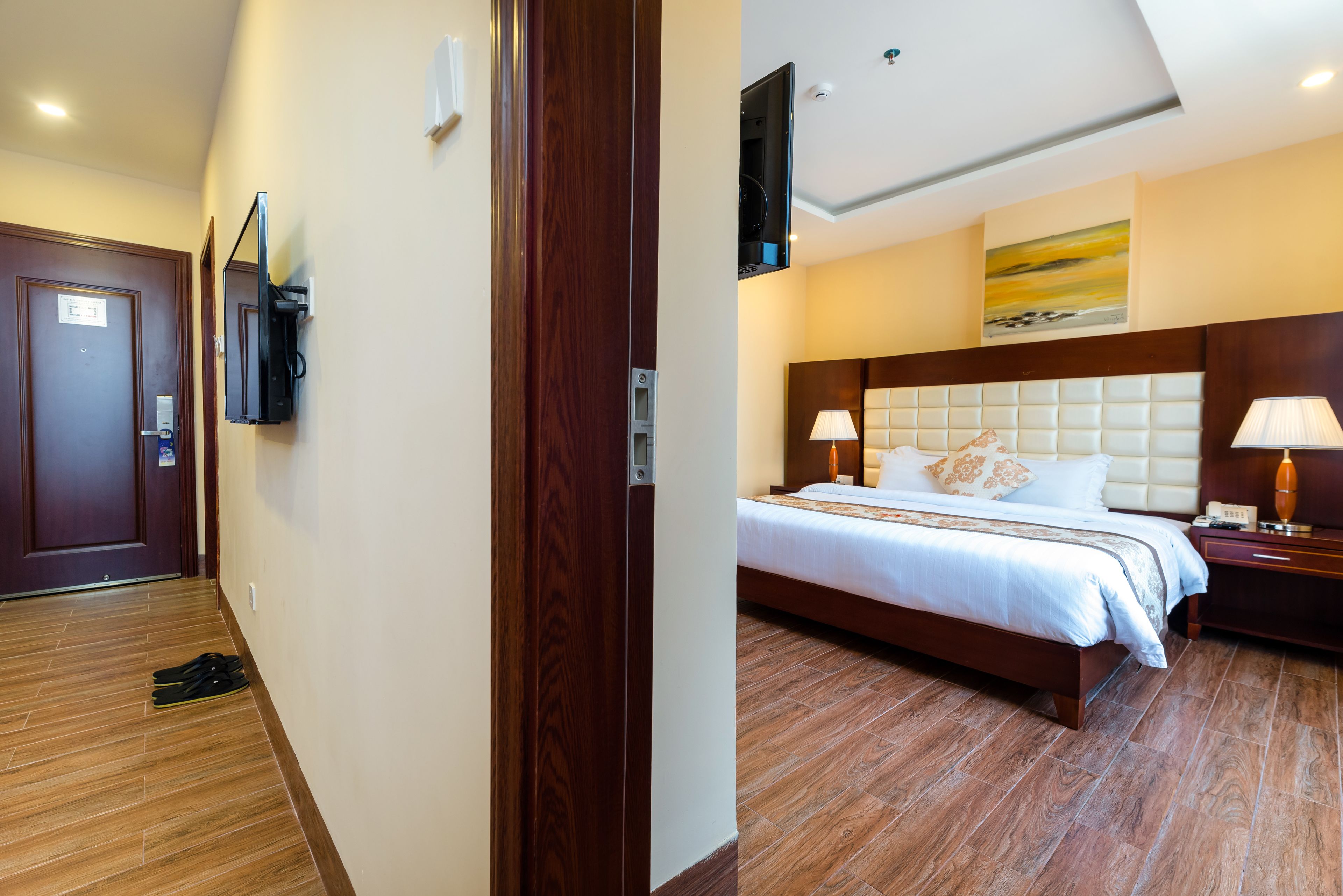 Quarto de hotel com piso de madeira, cama com roupa de cama branca, mesa de cabeceira com abajur e televisão fixada na parede.
