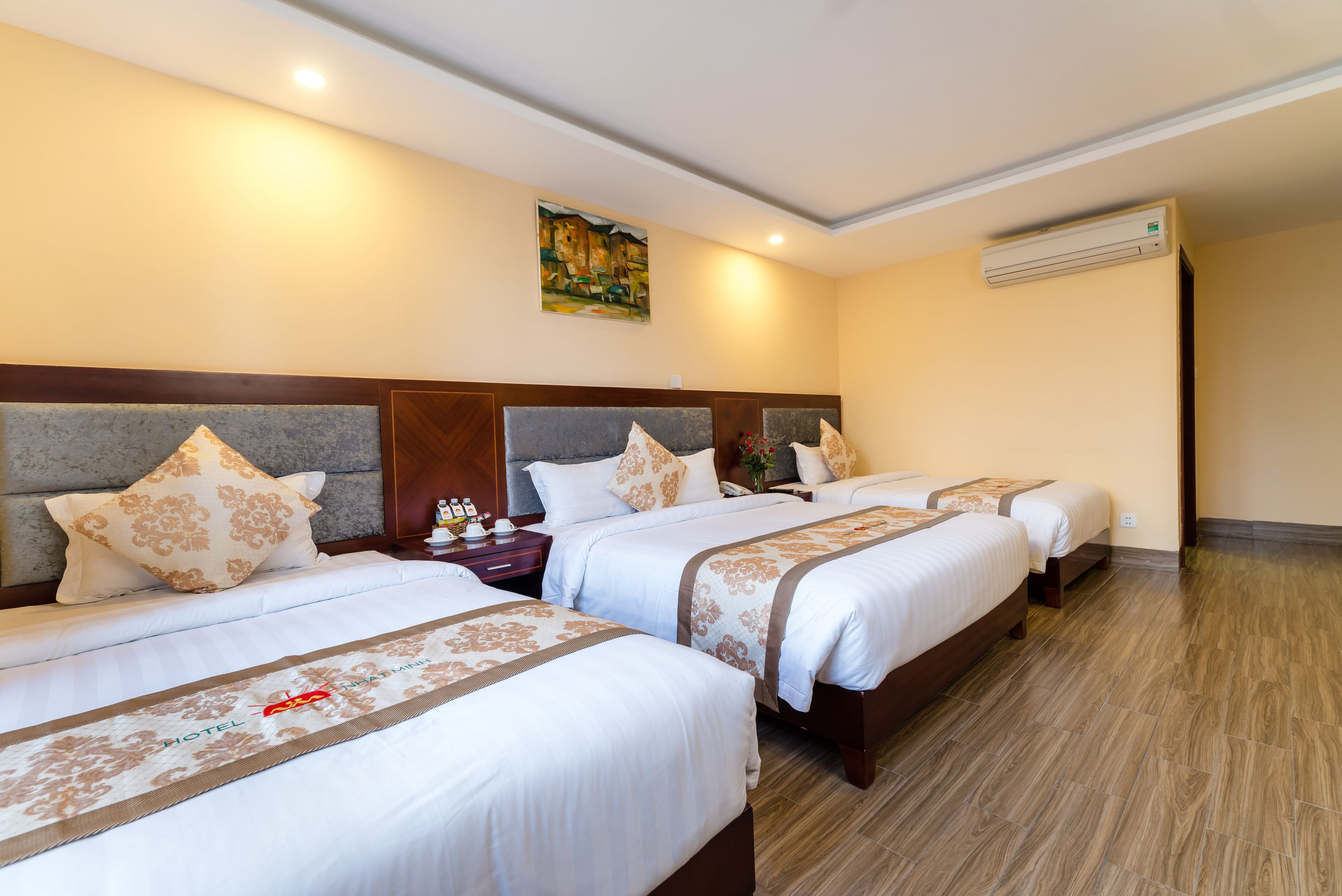 A hotel room with three beds, a headboard, a painting on the wall, and an air conditioning unit.